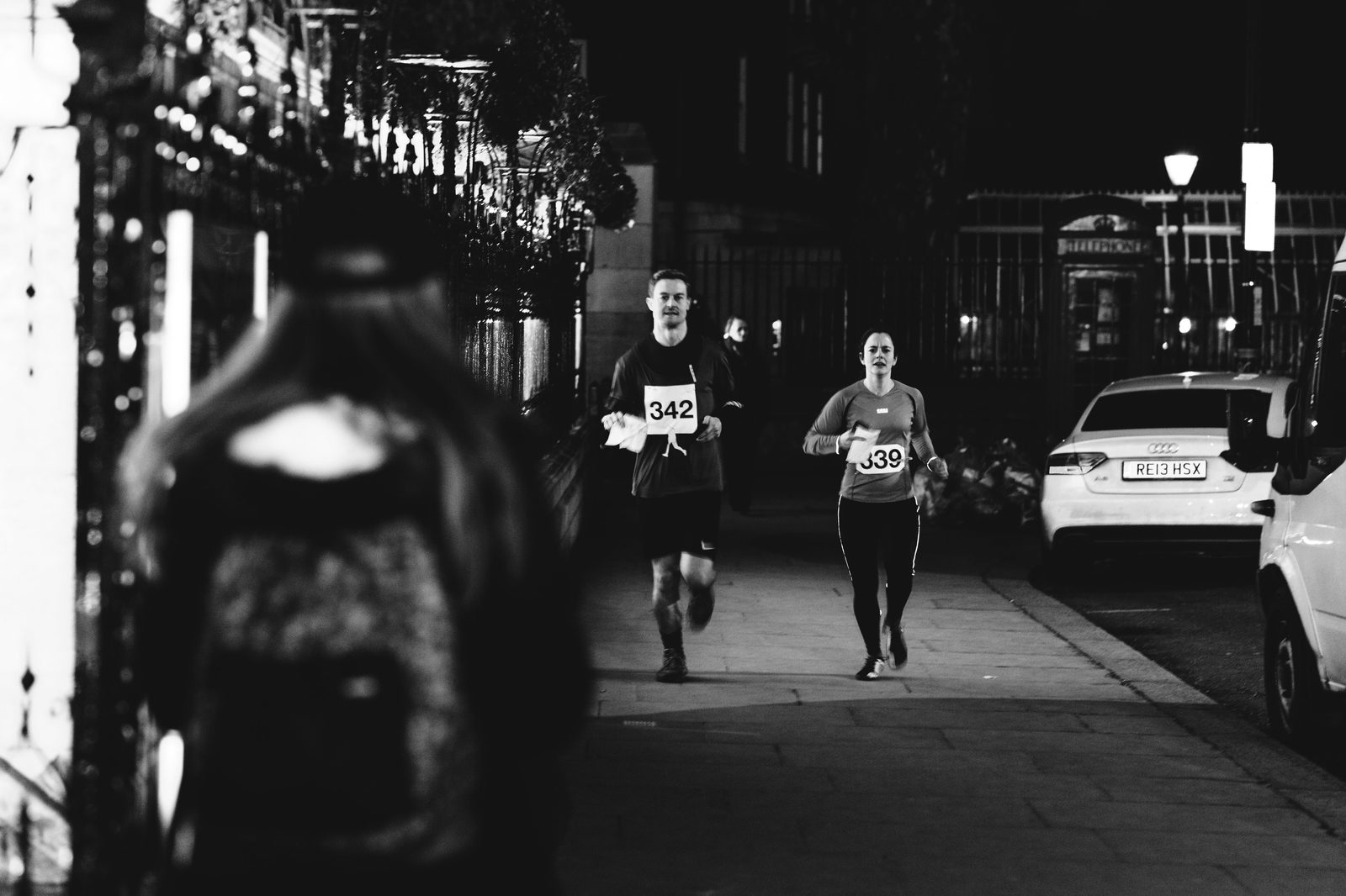 Black and white photo of two runners, wearing numbered bibs 342 and 339, running on a dimly lit city street at night. A person with a backpack walks in the foreground, with parked cars and city architecture visible in the background.
