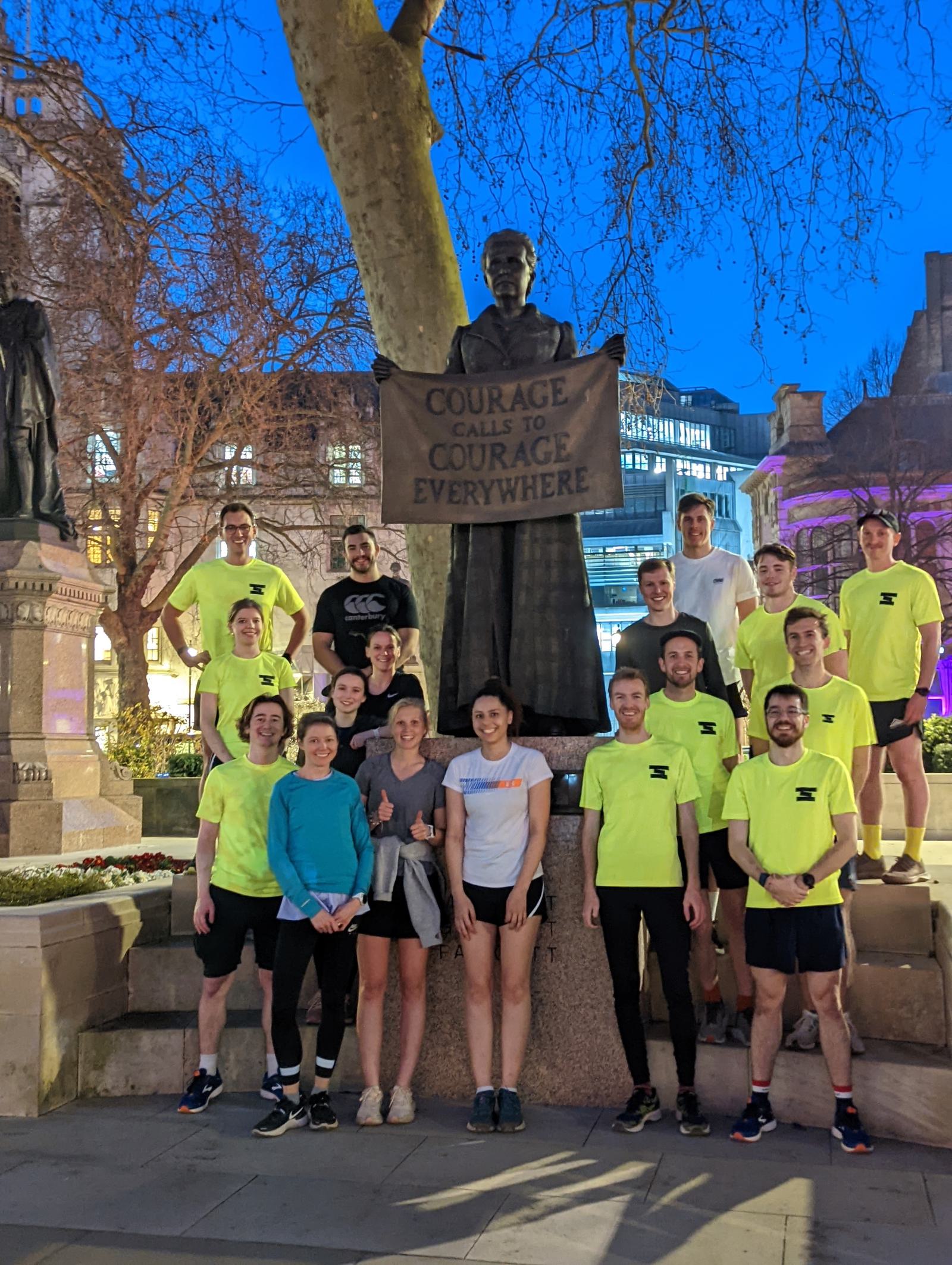 A group of people stands around a statue holding a sign that says, "Courage Calls to Courage Everywhere." Most of the group members are wearing bright yellow shirts and athletic gear, likely after a run, posing in a well-lit area with bare trees in the background.