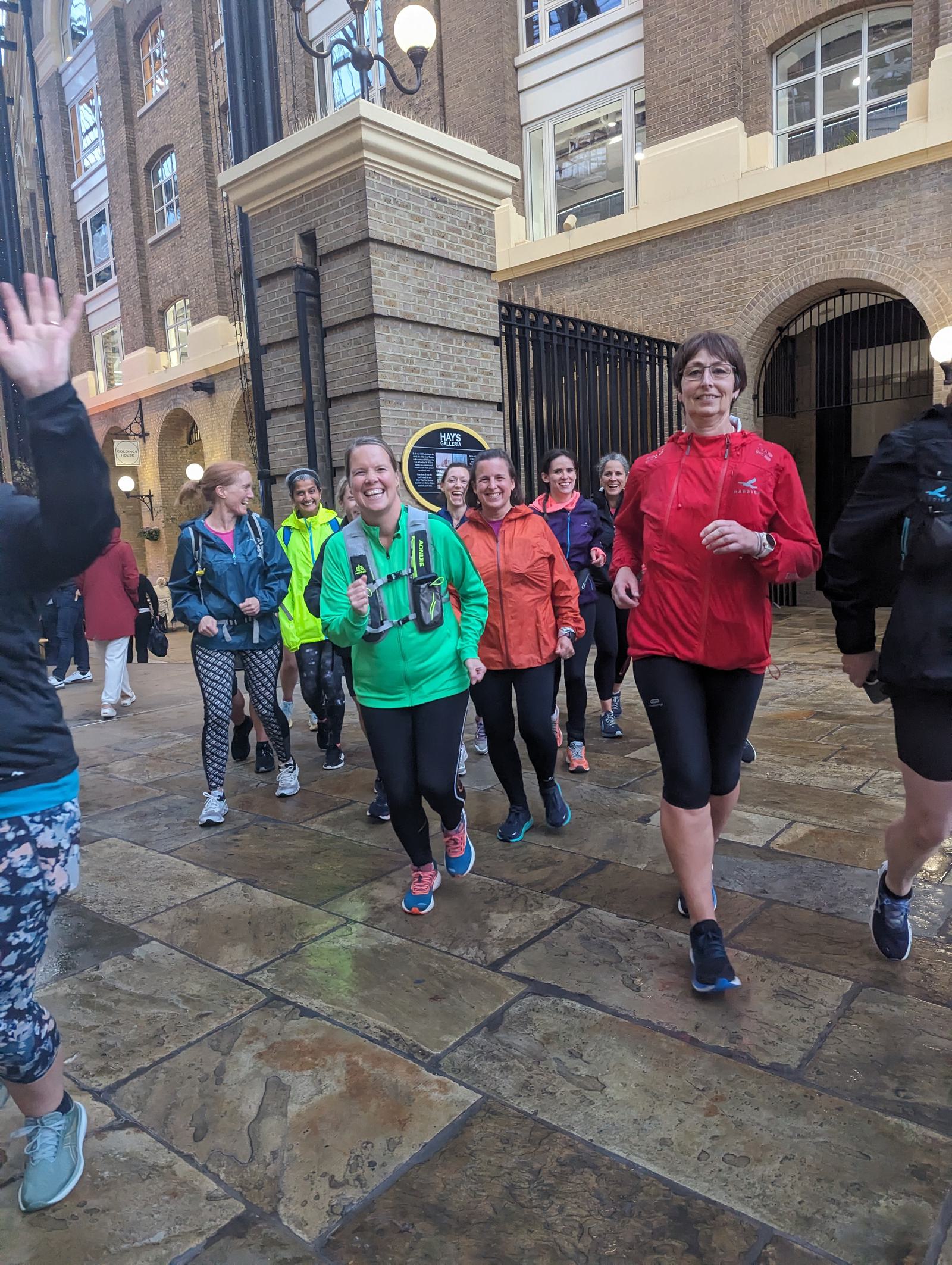 A group of women dressed in colorful sports attire are jogging through a cobblestone street lined with brick buildings. They appear cheerful and energetic, with some waving at the camera. The scene suggests a friendly and enjoyable group run.