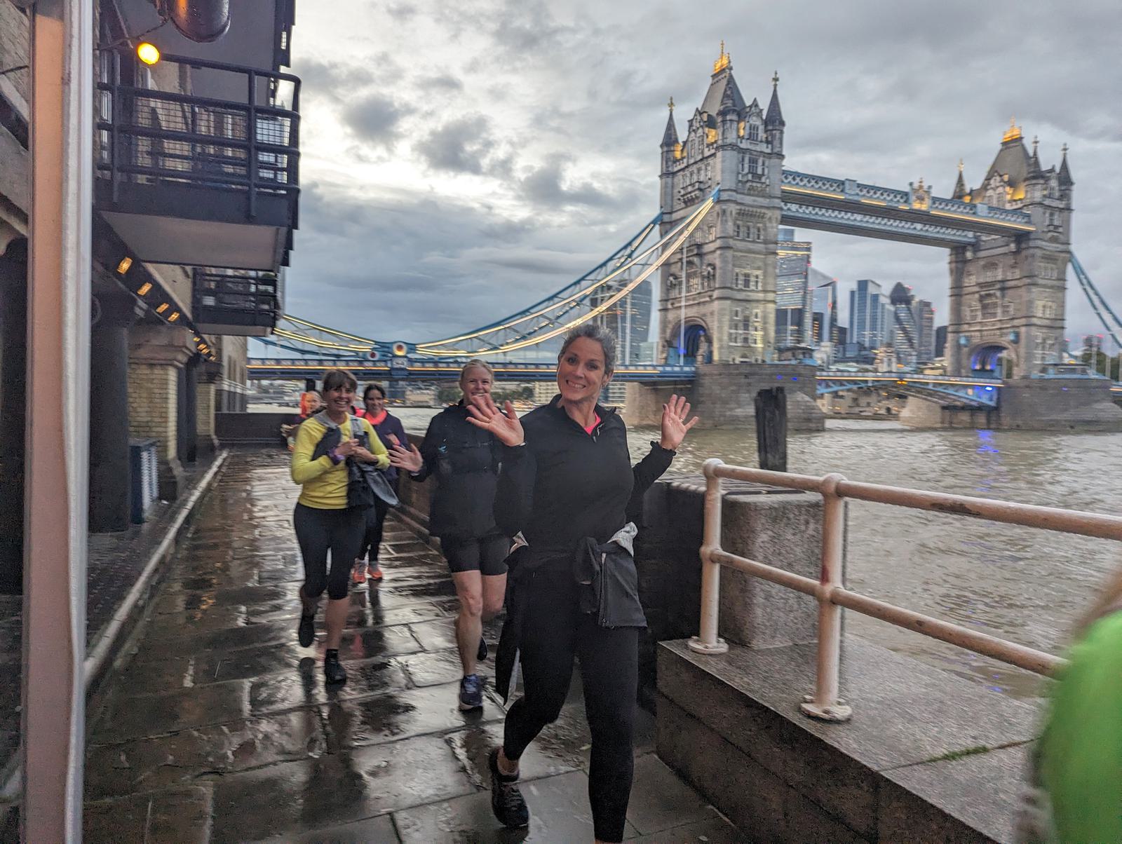 A group of smiling joggers wave as they run along a riverside path near Tower Bridge in London. The bridge's iconic towers are illuminated in the background, and the sky is overcast. The path is wet, indicating recent rain.