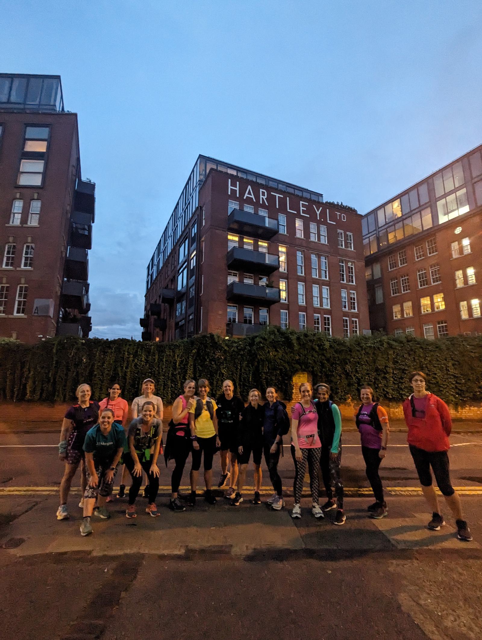 A group of people in athletic gear stands together on the sidewalk during dusk, posing in front of a brick building with the sign "HARTLEY LTD." The group appears ready for an outdoor or running activity, with some smiling at the camera and others looking relaxed.
