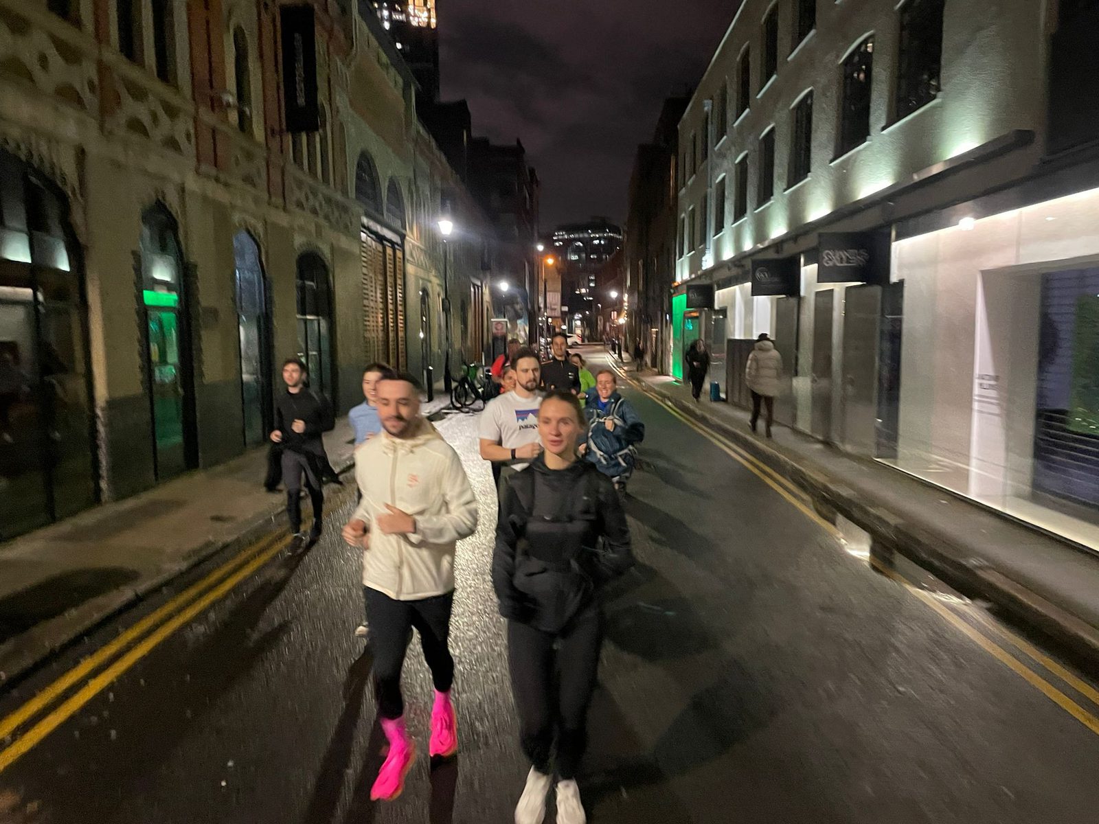 A group of people jogging on a dimly lit street at night. The street is flanked by modern buildings with illuminated storefronts and older, ornate brick buildings. Some pedestrians are also visible in the background. One person is wearing bright pink shoes.