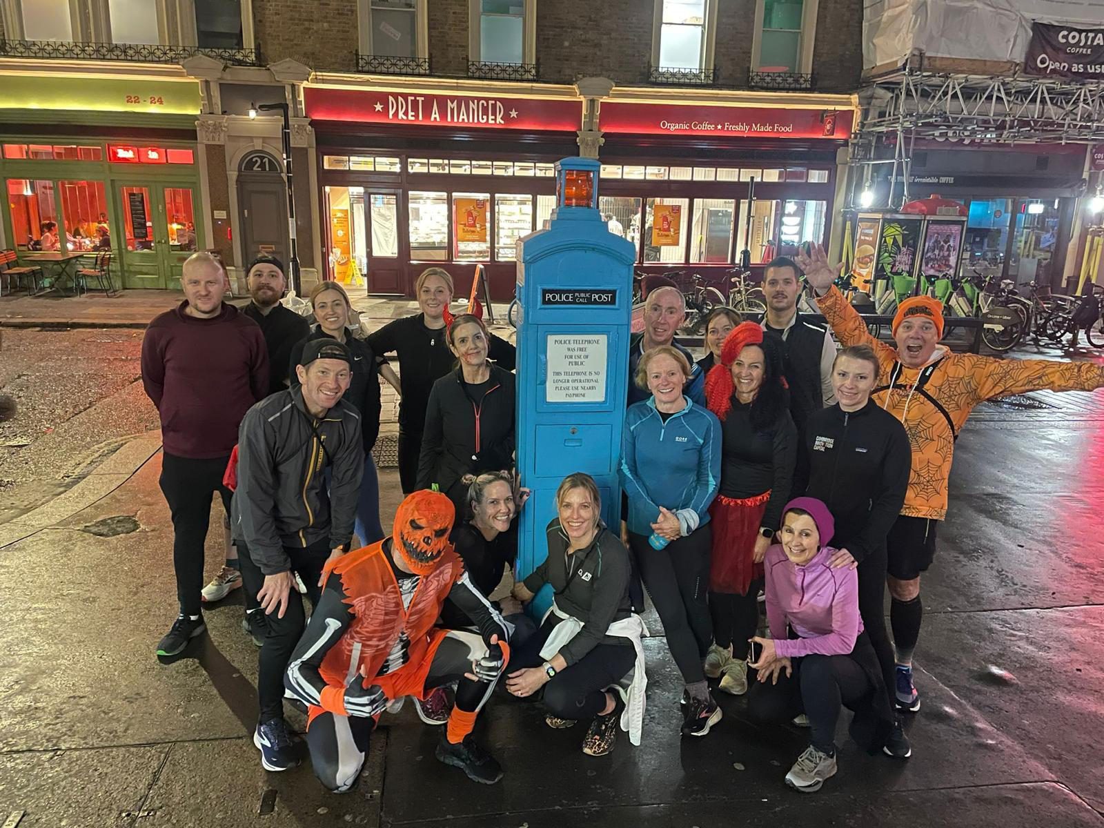 A group of people, some dressed in Halloween-themed costumes, stand together smiling in front of a blue police box on a nighttime city street. The background includes brightly lit storefronts, including a Pret A Manger restaurant.