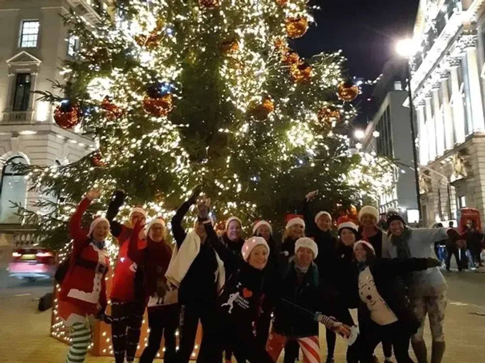 A group of people dressed in festive attire, including Santa hats and Christmas-themed clothing, stands in front of a large, ornately decorated Christmas tree adorned with lights and ornaments. They are posing and smiling happily on a city street at night.