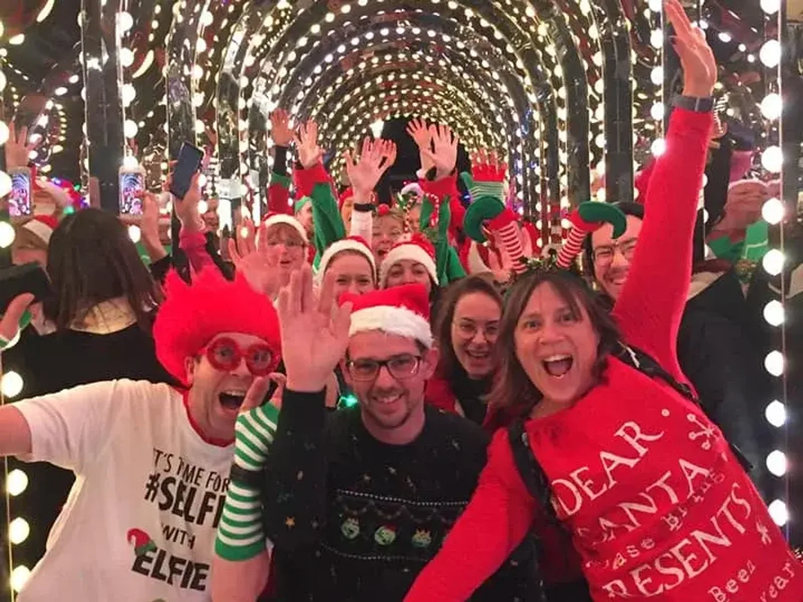 A group of cheerful people, some in festive sweaters, Santa hats, and holiday-themed costumes, pose happily in a tunnel decorated with strings of lights. They are smiling, waving, and celebrating the festive season.
