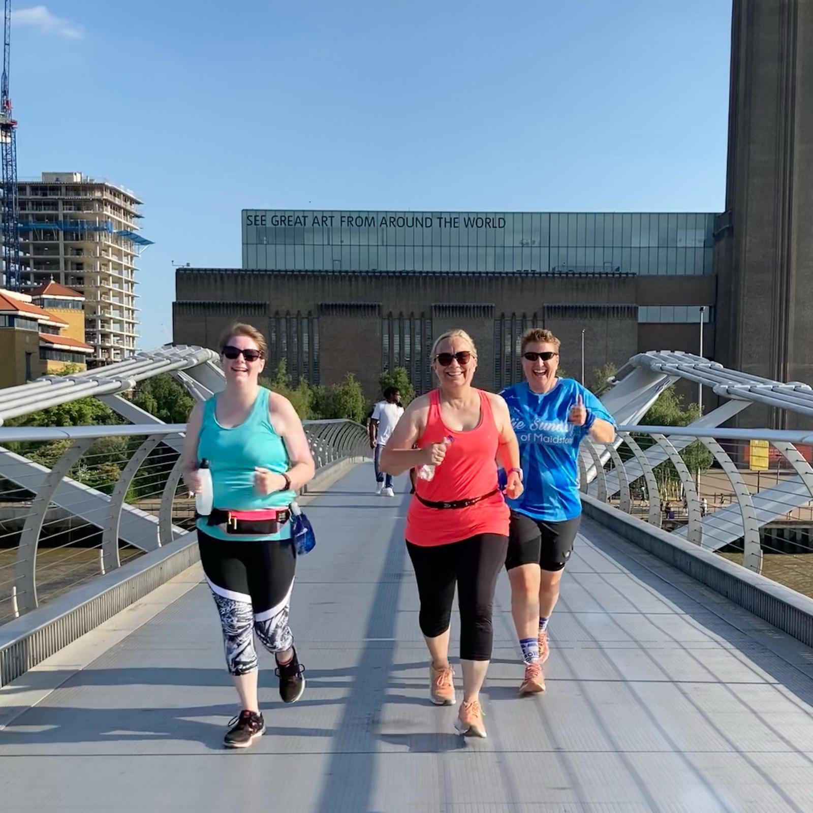 Three women are jogging on a modern footbridge in bright daylight with smiles on their faces. In the background, there is a tall industrial-style building with a sign that reads "SEE GREAT ART FROM AROUND THE WORLD." They are dressed in athletic wear.