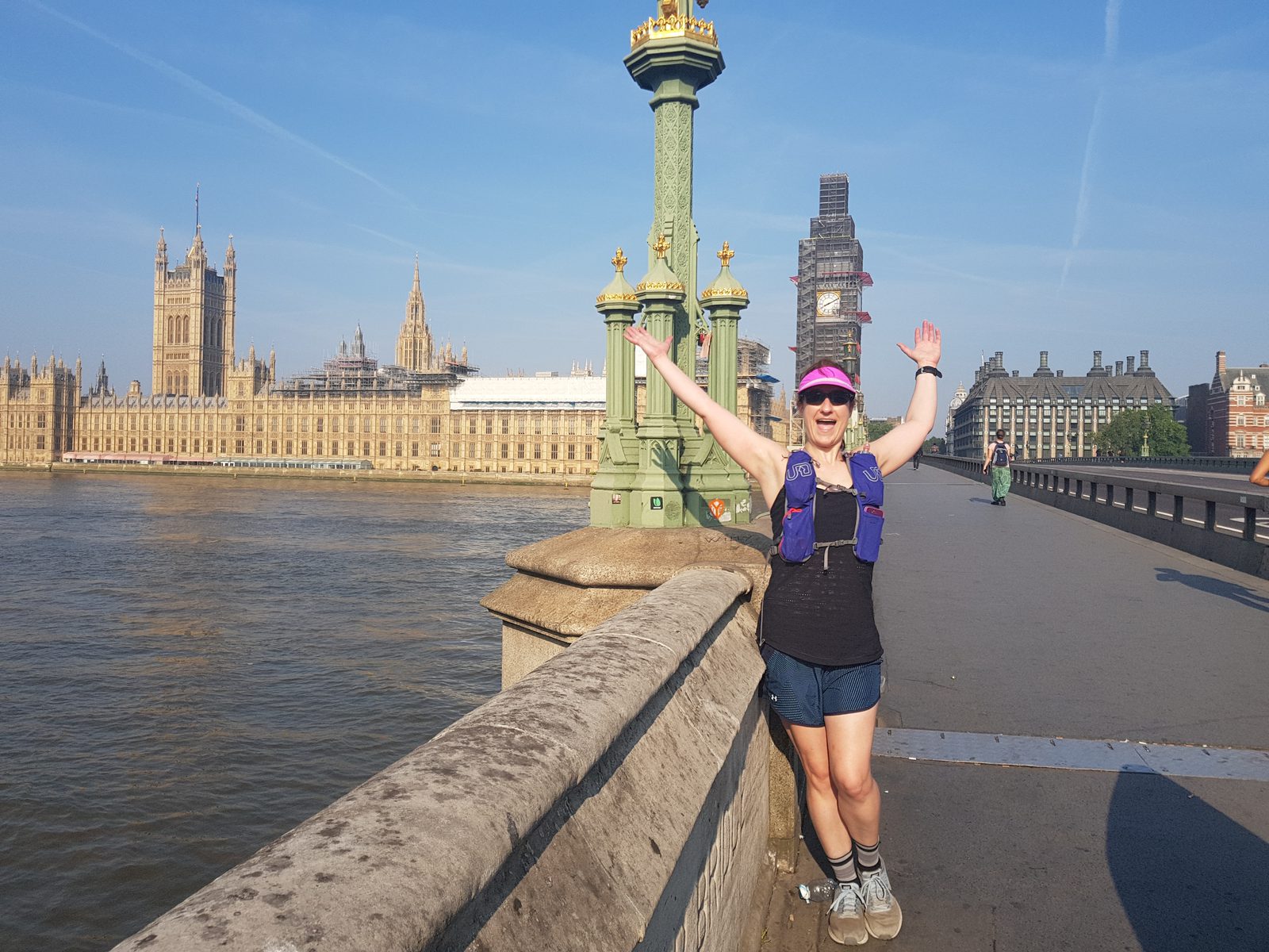 A person wearing a pink cap, sunglasses, and sporty attire stands on a bridge with their arms raised in joy. The background features the River Thames and the iconic Houses of Parliament and Big Ben in London on a sunny day.