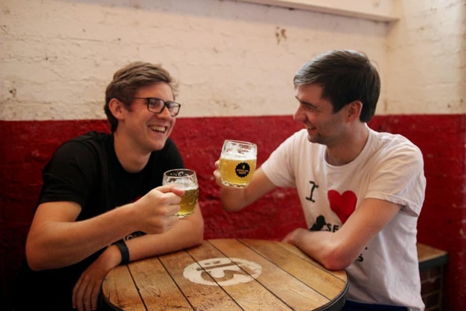 Two young men sitting at a wooden table in a bar, smiling and clinking their beer glasses. One man is wearing glasses and a black shirt, while the other is wearing a white shirt with text on it. The table has the number '13' painted on it. Red and white wall in the background.