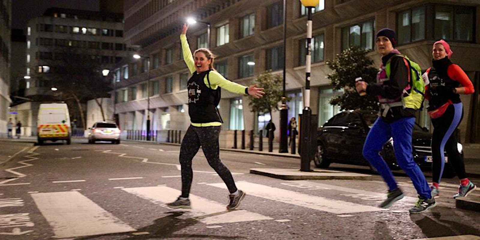 Three runners in colorful athletic wear cross a crosswalk at night on a city street. A woman in the center waves energetically with both arms raised while the other two follow. Buildings, streetlights, and a van are visible in the background.
