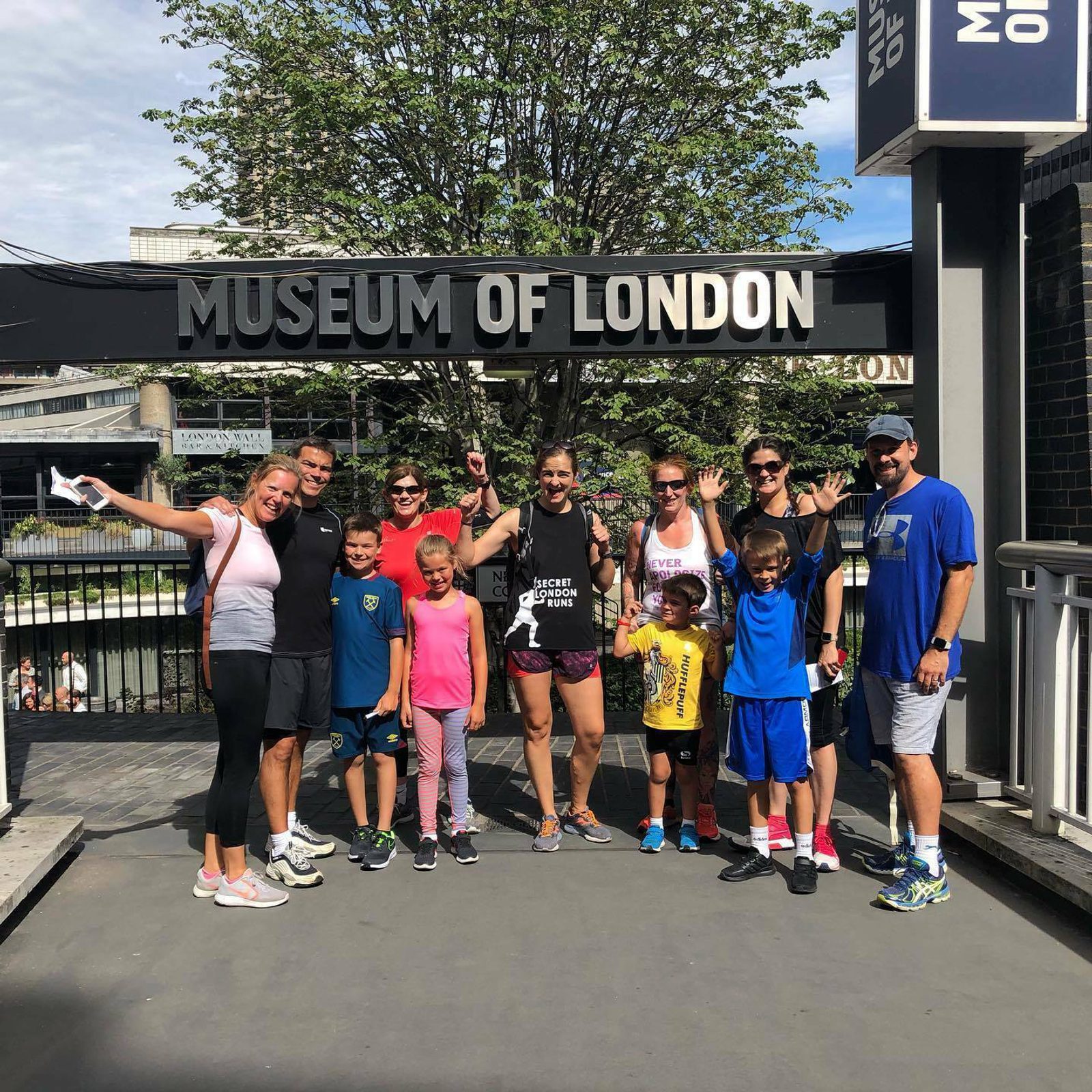 A group of adults and children standing in front of the "Museum of London" sign. They are smiling, waving, and posing for the camera. The weather is sunny, and there are trees and buildings in the background. The group appears to be enjoying their visit.