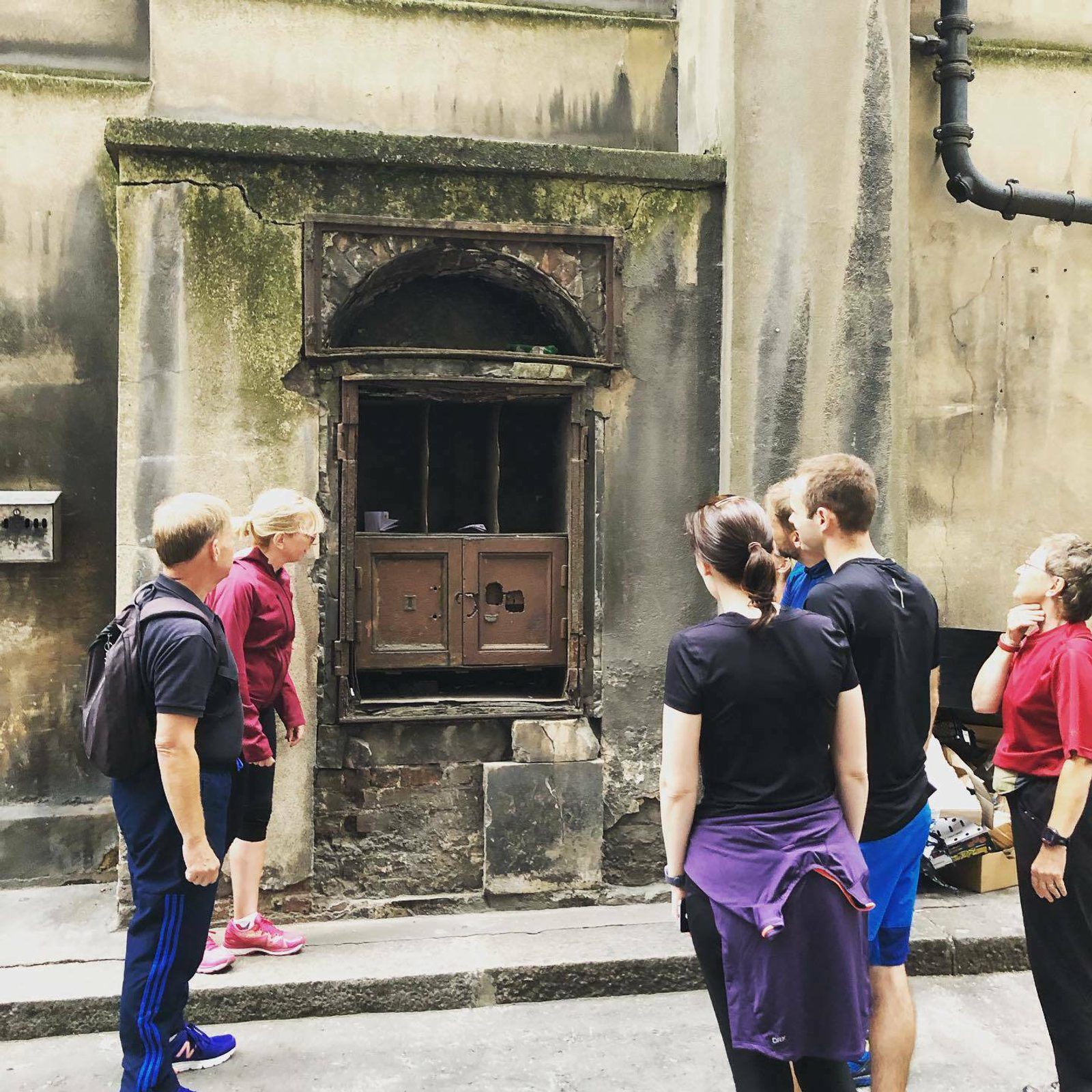 A group of six people stands in front of an old, weathered wall with a small, arched wooden door. The wall is aged and has visible cracks and moss. Some of the individuals appear to be listening to a guide or discussing the structure.