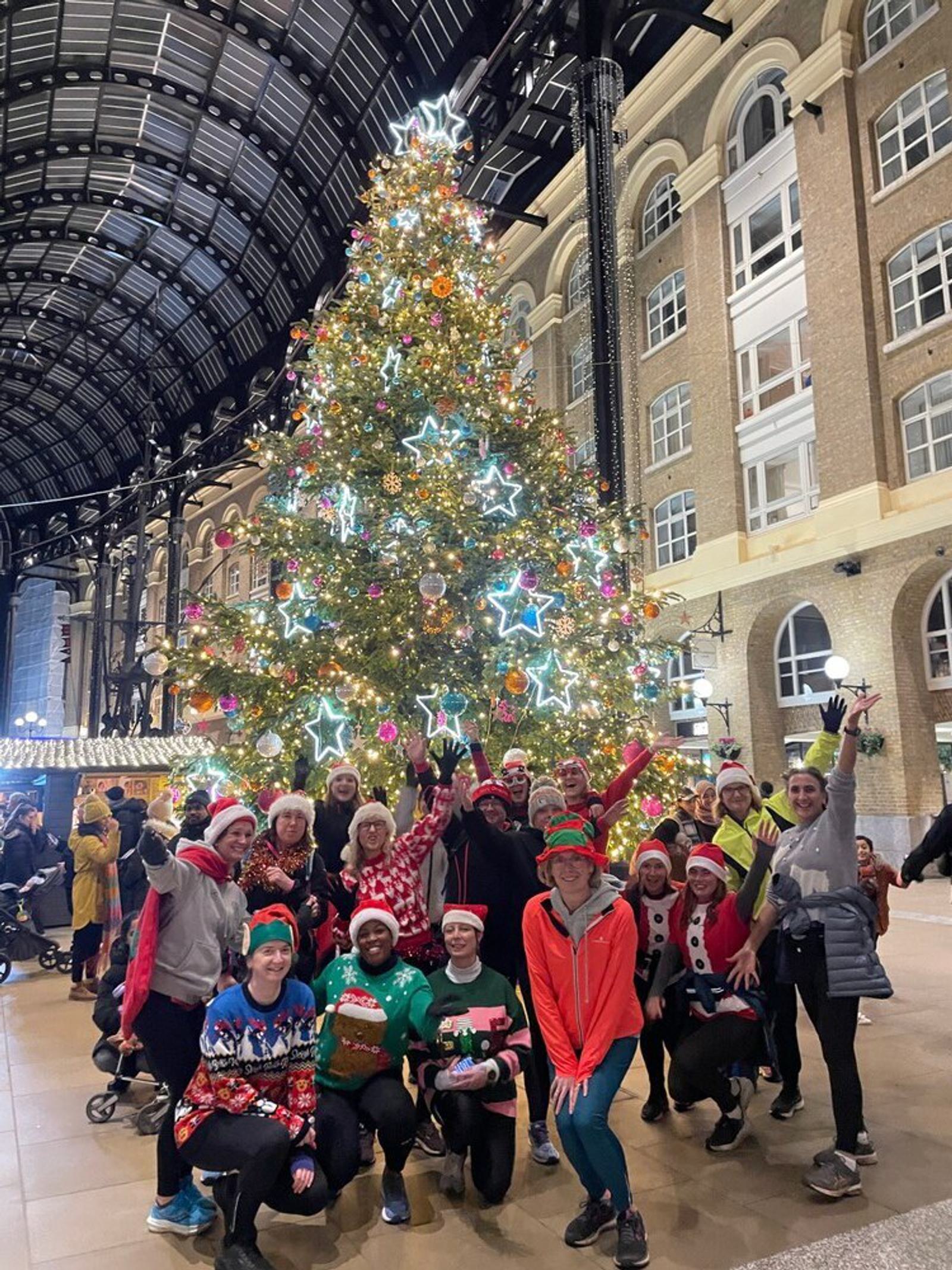 A group of people in festive attire, including Santa hats and holiday-themed sweaters, pose in front of a large, decorated Christmas tree with colorful lights and ornaments. They are inside a spacious, high-ceilinged building adorned with large windows.