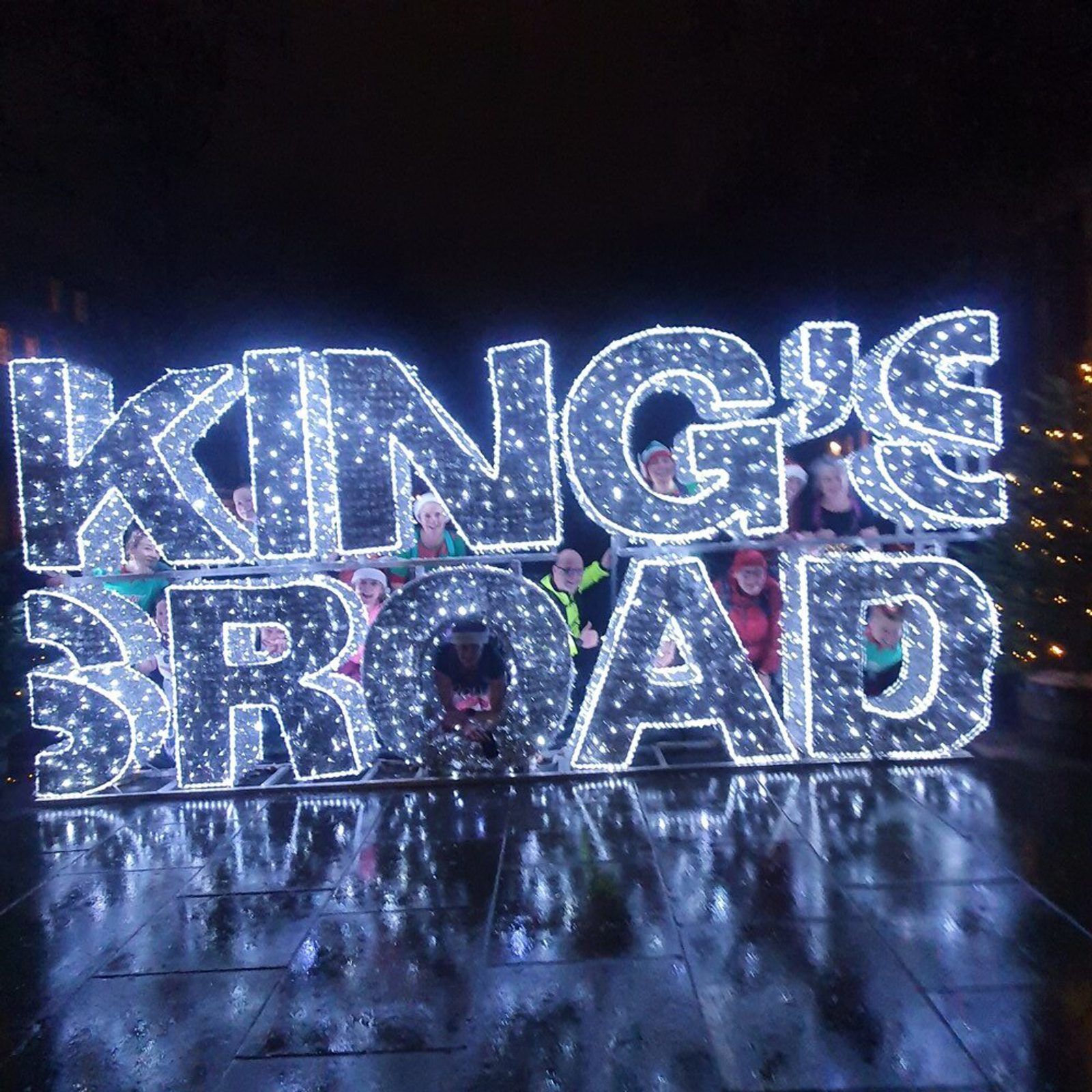 A group of people stands behind and within a large illuminated sign that reads "KING'S ROAD" in bright white lights. The ground is wet, reflecting the light, and it appears to be nighttime. The festive atmosphere suggests a holiday or celebratory event.