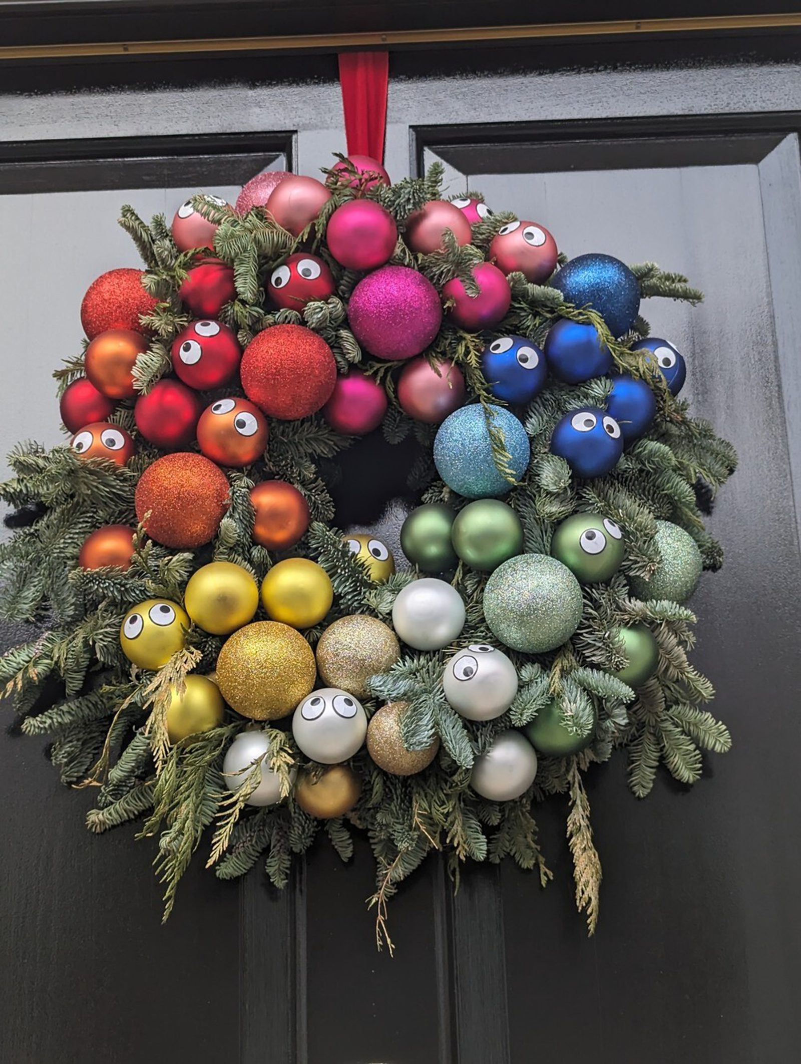 A festive wreath decorated with colorful Christmas ornaments, each with googly eyes, creating a playful, almost cartoonish effect. The ornaments vary in color from green, blue, and purple to red, orange, and gold, all attached to a green evergreen base.
