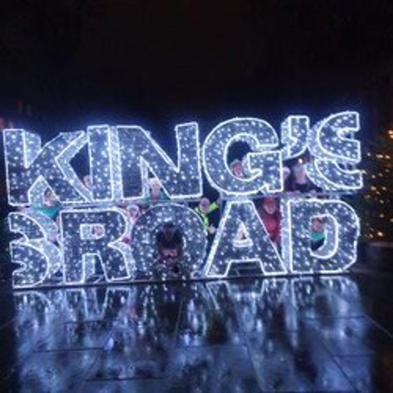 Illuminated sign with the words "KING'S ROAD" in large, bright lights on a reflective surface. The display is set against a dark background, creating a festive atmosphere.