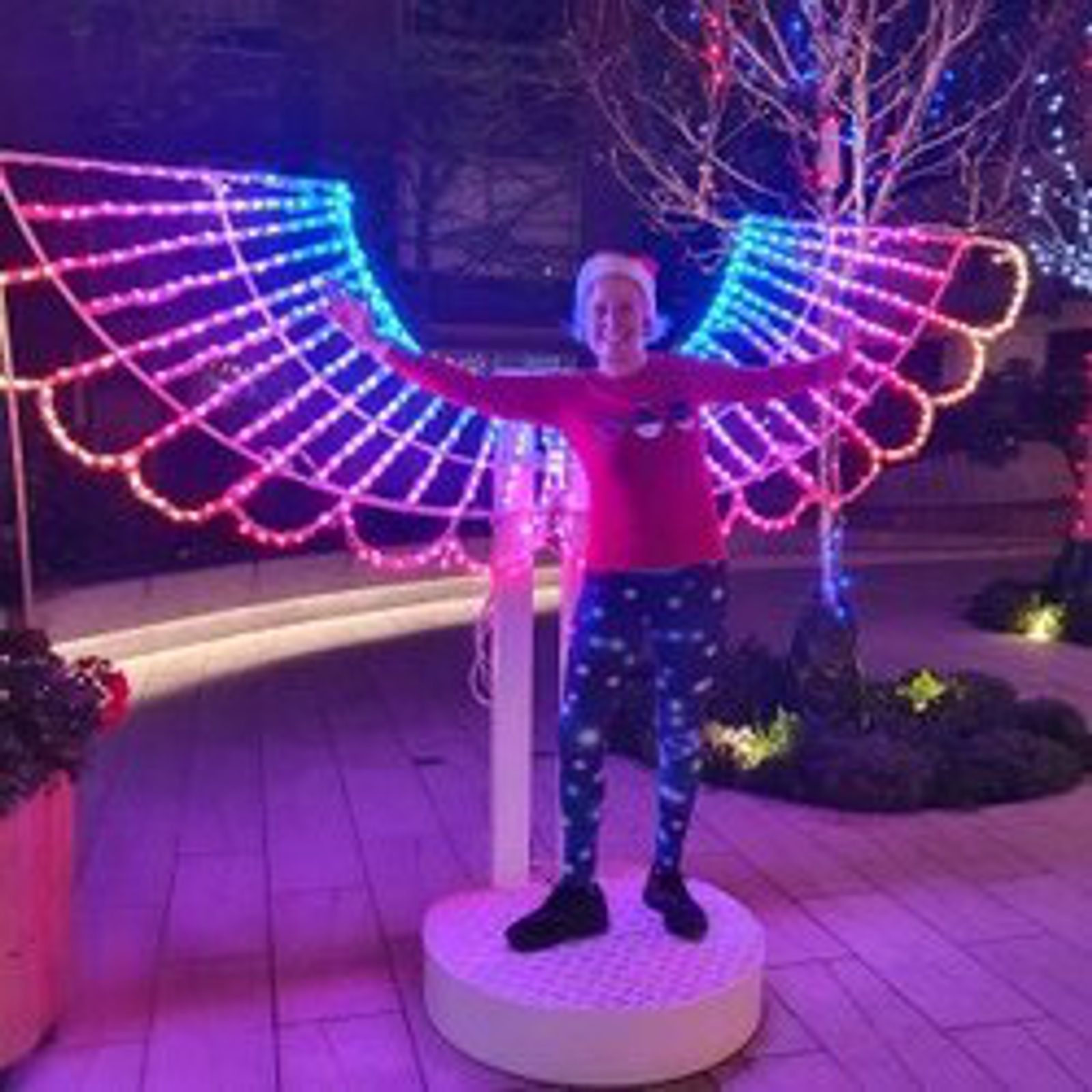 A person is standing on a circular platform, posing with outstretched arms in front of colorful, illuminated wings. The wings are outlined with bright lights in shades of pink, blue, and purple. The scene appears to be in an outdoor, festive setting at night.