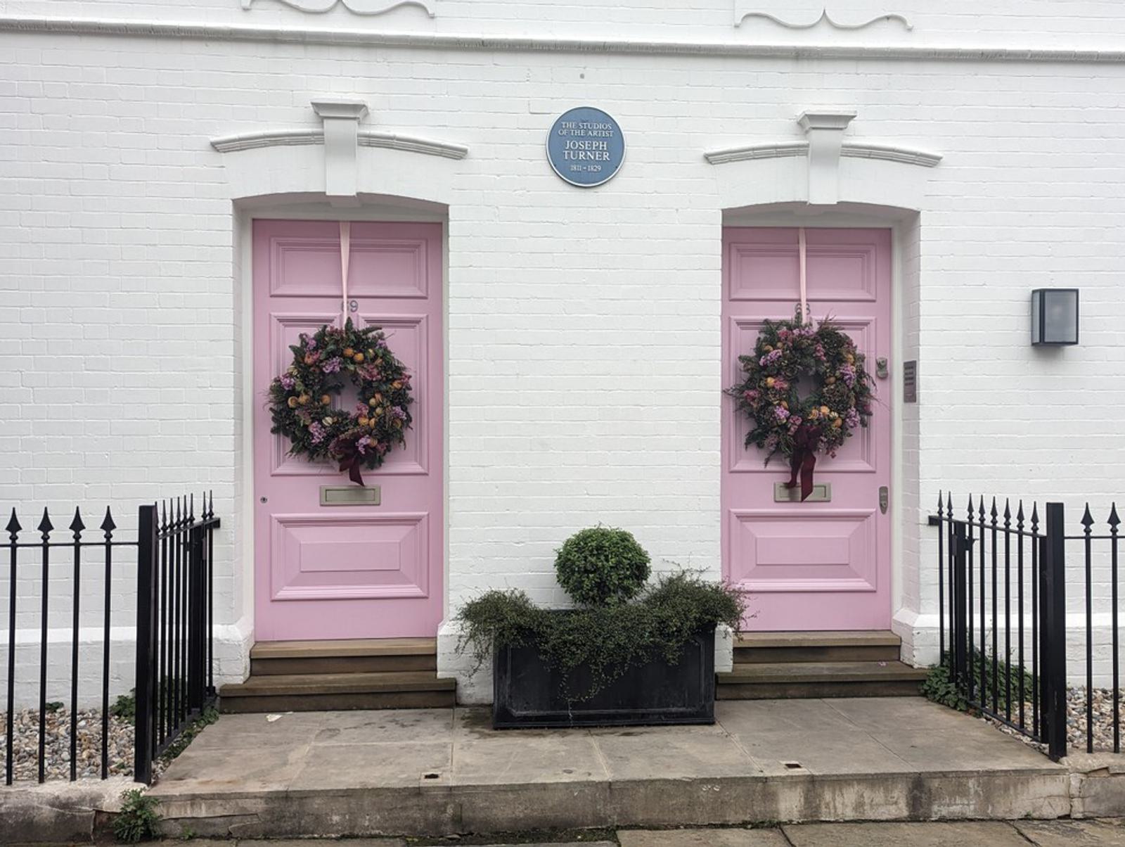 Two adjacent pastel pink doors, each adorned with a festive wreath, set against a white brick wall. A black metal fence runs along the front, with a potted greenery between the doors. A blue circular plaque is mounted above the left door.