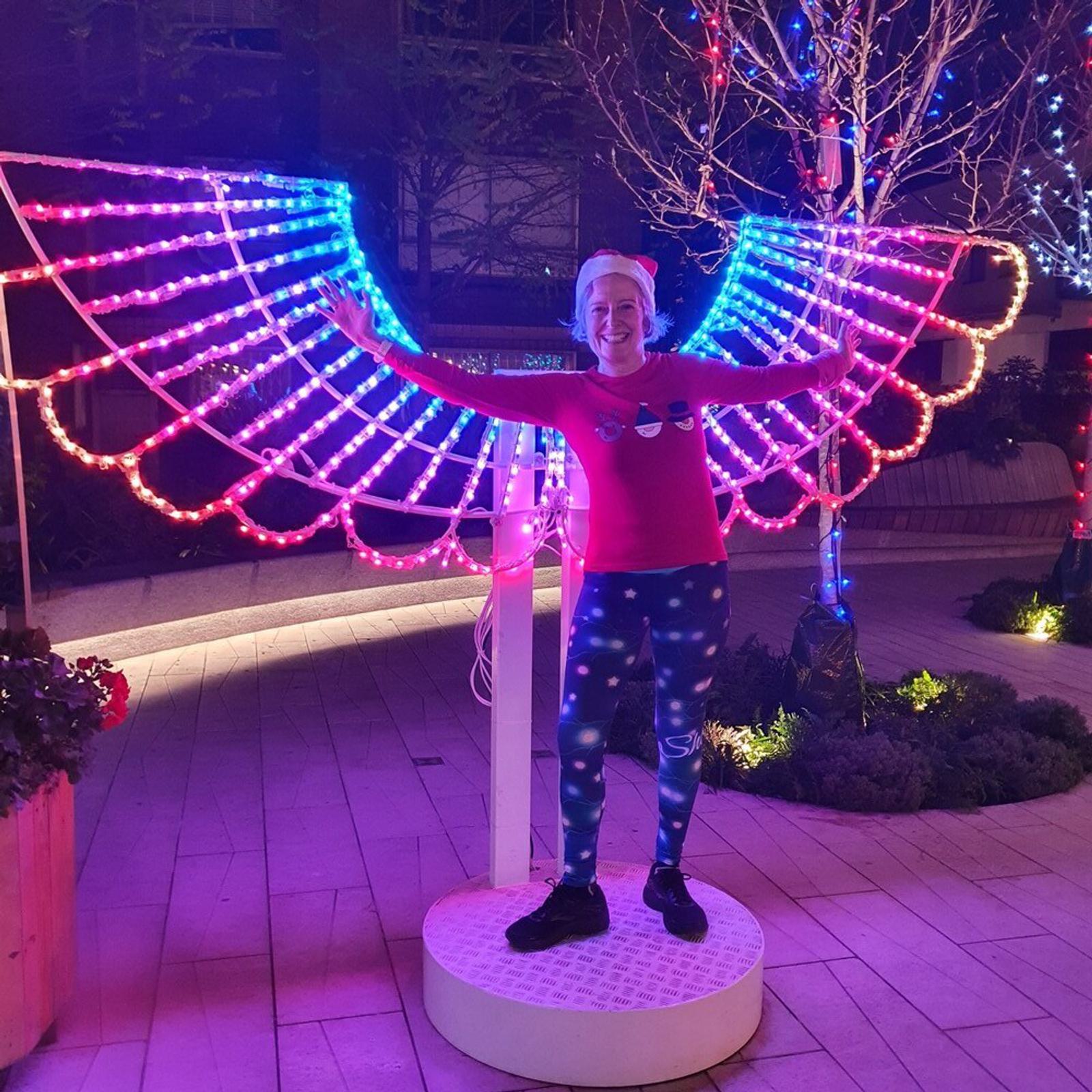 A person wearing a festive sweater and galaxy print leggings poses in front of a neon light installation shaped like wings. The wings glow in vibrant hues of pink, blue, and purple, creating an angelic effect. Trees with lights decorate the background.