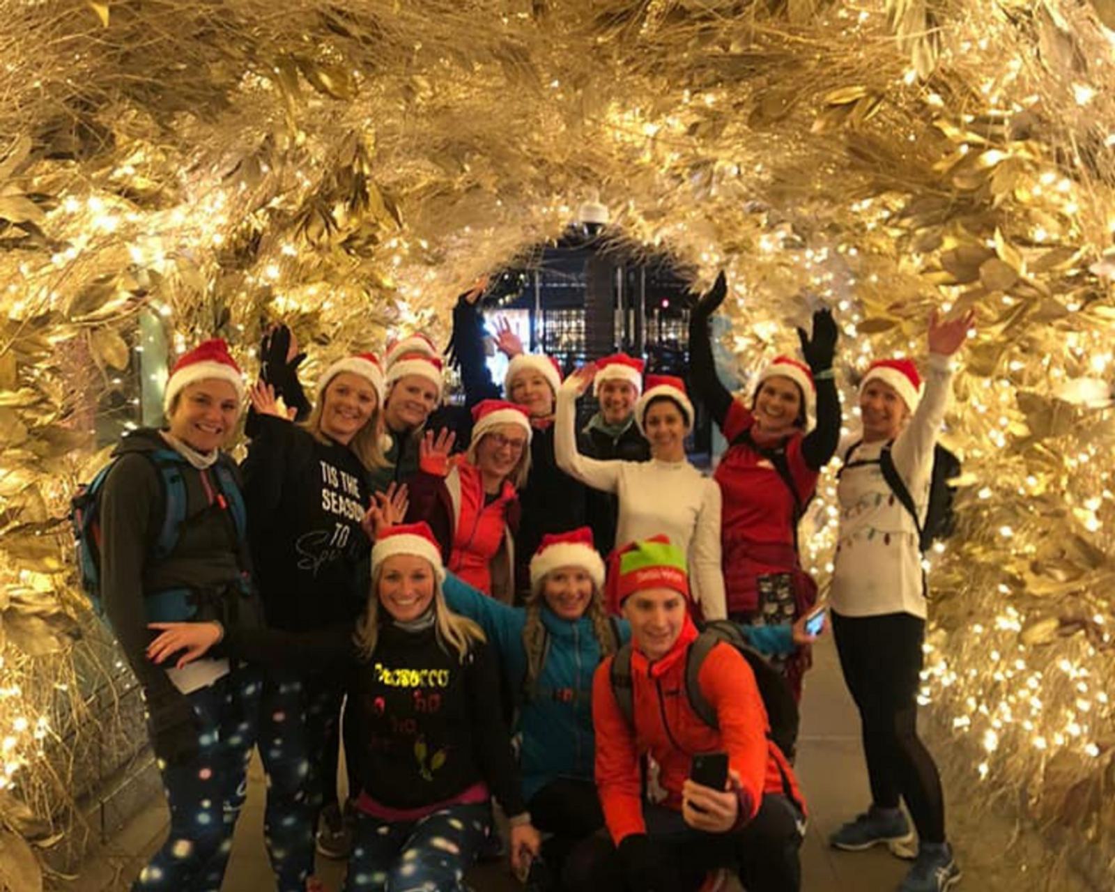 A group of thirteen people wearing Santa hats pose for a photo under a tunnel of golden holiday lights. They are smiling and waving at the camera, dressed in festive attire, including Christmas-themed leggings and sweaters. The scene is cheerful and festive.