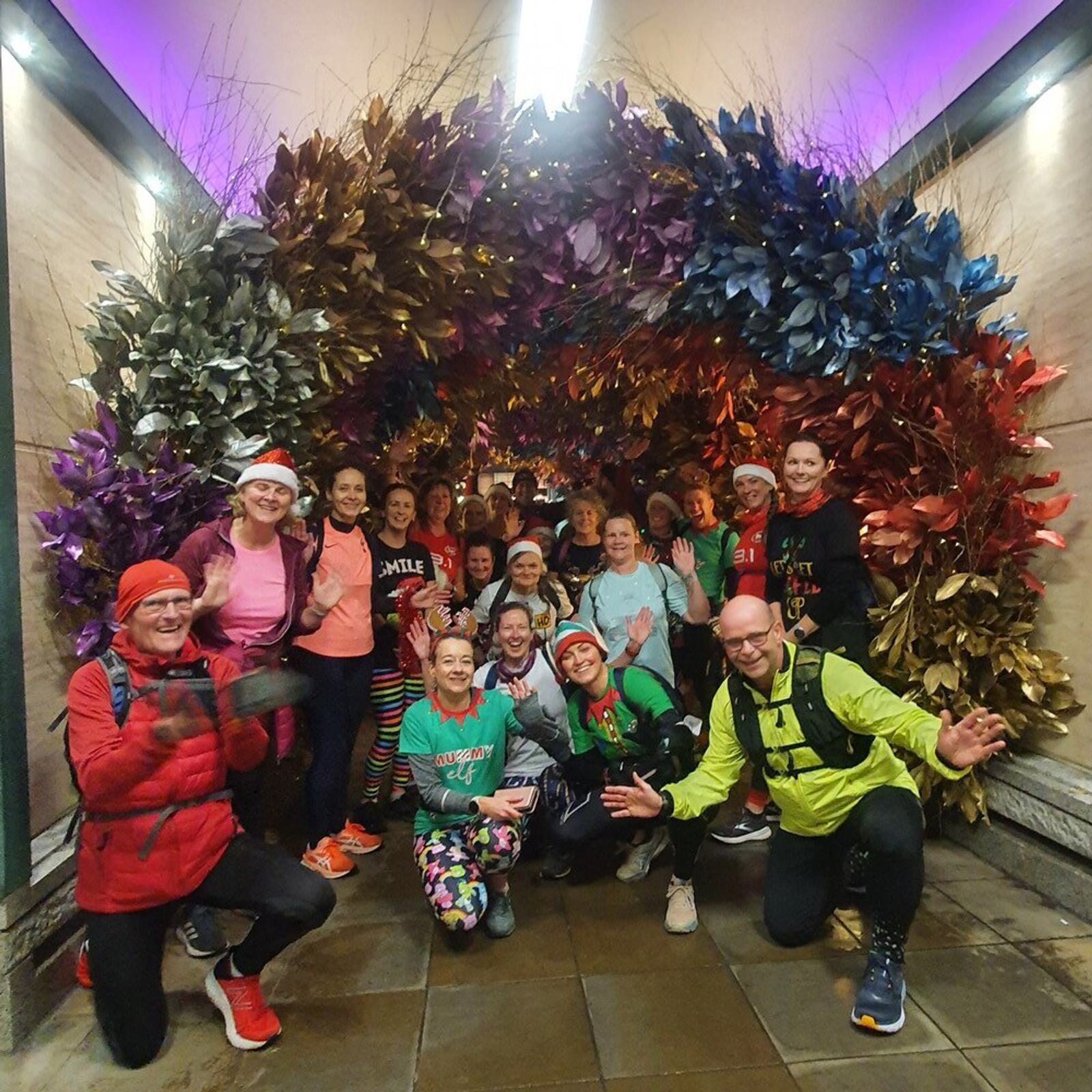 A group of people in colorful athletic clothing pose for a photo in front of a festive arch decorated with vibrant, metallic-colored leaves in shades of gold, silver, pink, and blue. Some are smiling and waving at the camera, and the setting appears to be indoors.