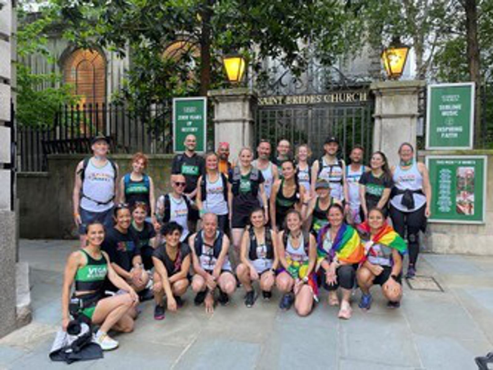 A group of people in athletic wear pose in front of a church with a sign that reads "St. Bride's Church." Some individuals wear colorful pride flags. Trees and church signs are in the background.