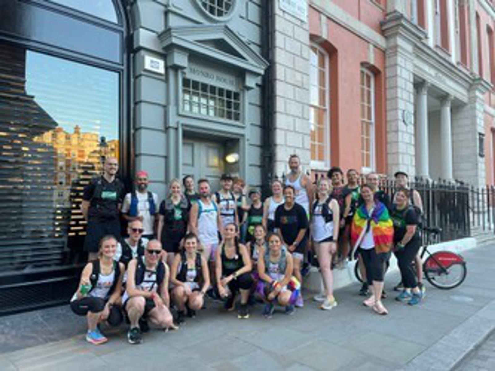 A group of people in athletic wear poses happily in front of a historic building with large windows and a street lamp. Some wear running gear and backpacks, one person has a rainbow flag, and a bicycle is partially visible on the right.