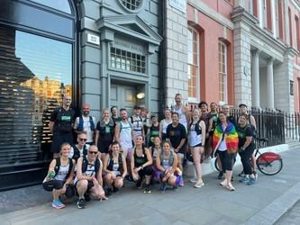 A group of people in athletic wear poses happily in front of a historic building with large windows and a street lamp. Some wear running gear and backpacks, one person has a rainbow flag, and a bicycle is partially visible on the right.
