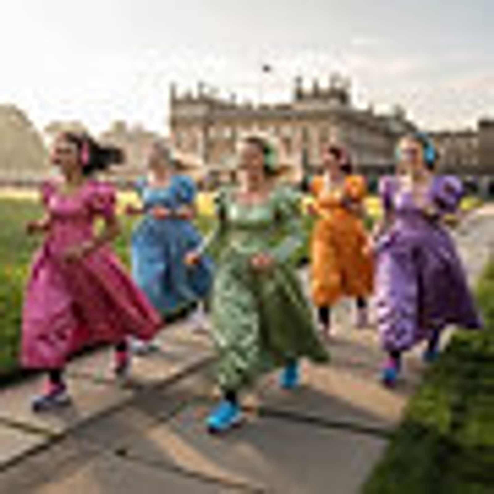 Five women in colorful Regency-style dresses walk and smile outside a grand historic building, with green lawns and sunlight in the background.