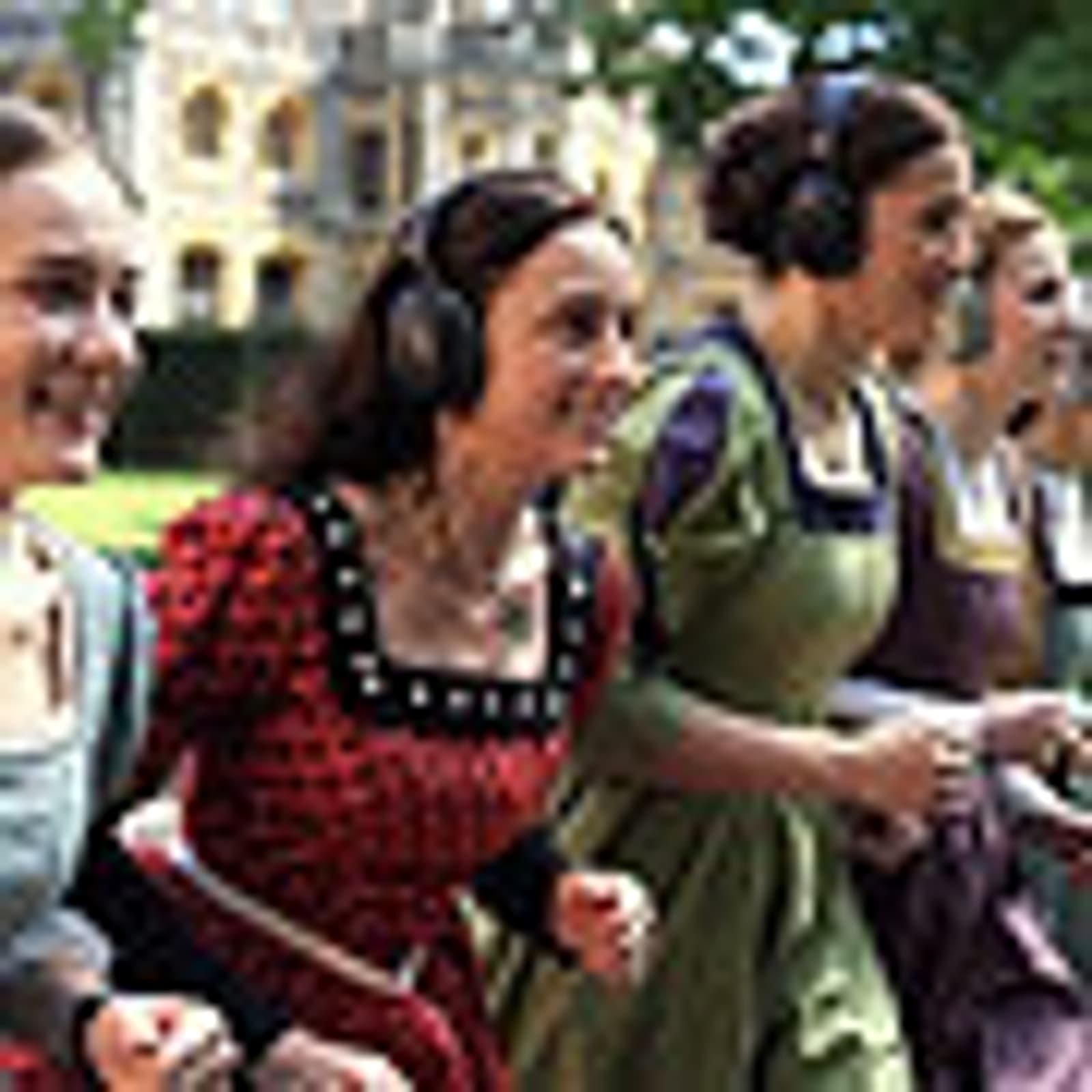 Four women in Renaissance-style dresses, wearing headphones, smiling and walking together outdoors, with historic buildings and greenery in the background.