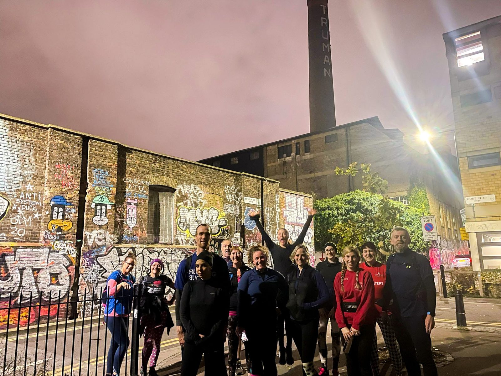 A group of people stand together at night in an urban area with graffiti-covered walls. The background features a tall brick chimney with "TRUMAN" written on it. The group is casually dressed, suggesting they might be out for a walk or run.