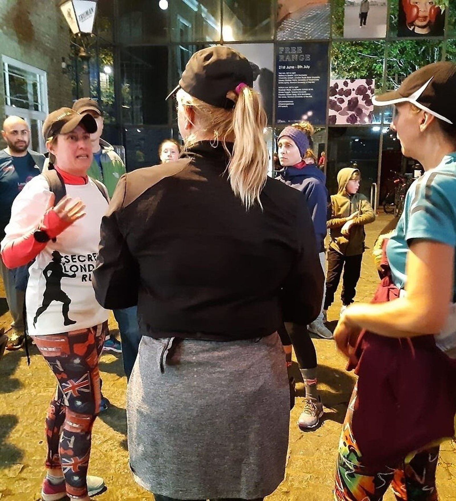 A group of people in athletic attire, including leggings and caps, stand outside a building at night, engaging in a conversation. One person faces away from the camera, while others are visible from the front and sides. Some people are wearing layers to keep warm.