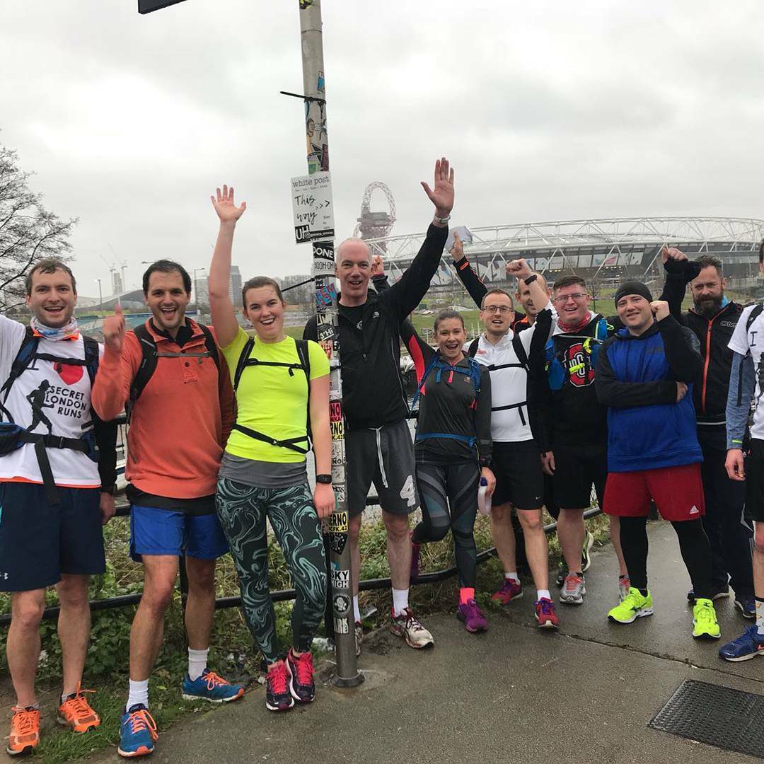 A group of ten runners, dressed in athletic gear, pose together outdoors near a pole covered in stickers. Some have their arms raised in celebration. In the background, a large stadium and an overcast sky are visible. The ground is wet, suggesting recent rain.