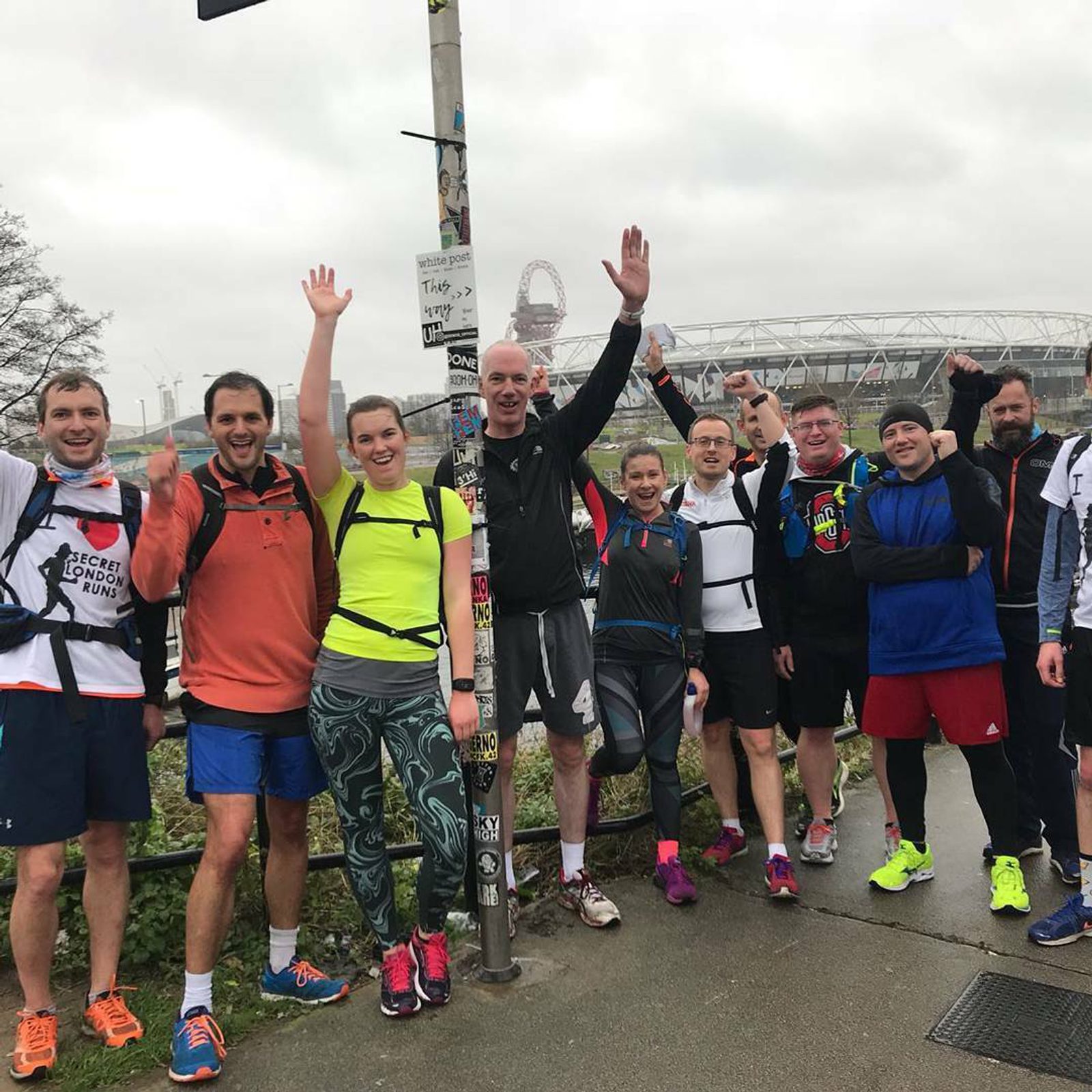 A group of ten runners, dressed in athletic gear, pose together outdoors near a pole covered in stickers. Some have their arms raised in celebration. In the background, a large stadium and an overcast sky are visible. The ground is wet, suggesting recent rain.
