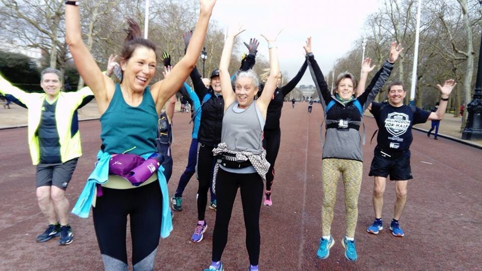 A group of runners is jumping joyfully with their arms raised on a park pathway. They appear to be in high spirits, wearing athletic clothing, and the background features trees and a cloudy sky. Some of the runners have fanny packs and are smiling.