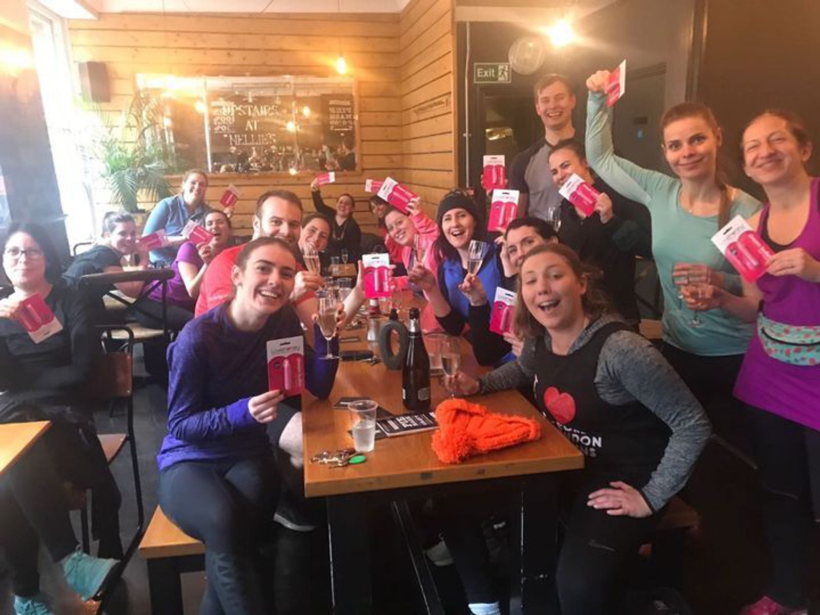 A group of people are sitting and standing around tables in a lively, indoor setting, smiling and holding up pink gift cards. They are dressed in casual athletic attire, and the background includes a wooden wall with writing and decorations.
