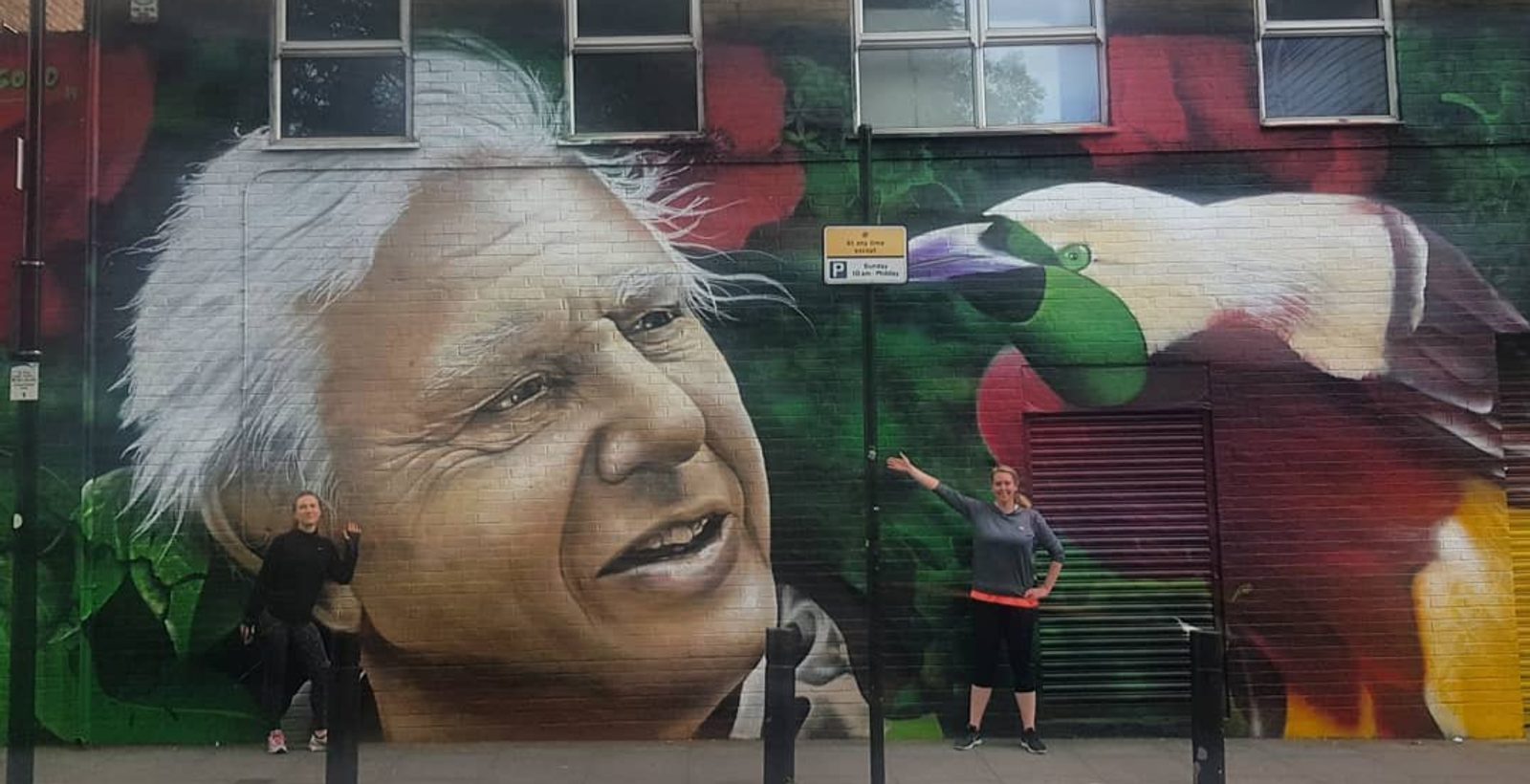 Two people pose in front of a large mural on a brick wall. The mural features a detailed portrait of an older man with white hair and a green toucan perched near his shoulder. The individuals are pointing towards the mural with smiles on their faces.