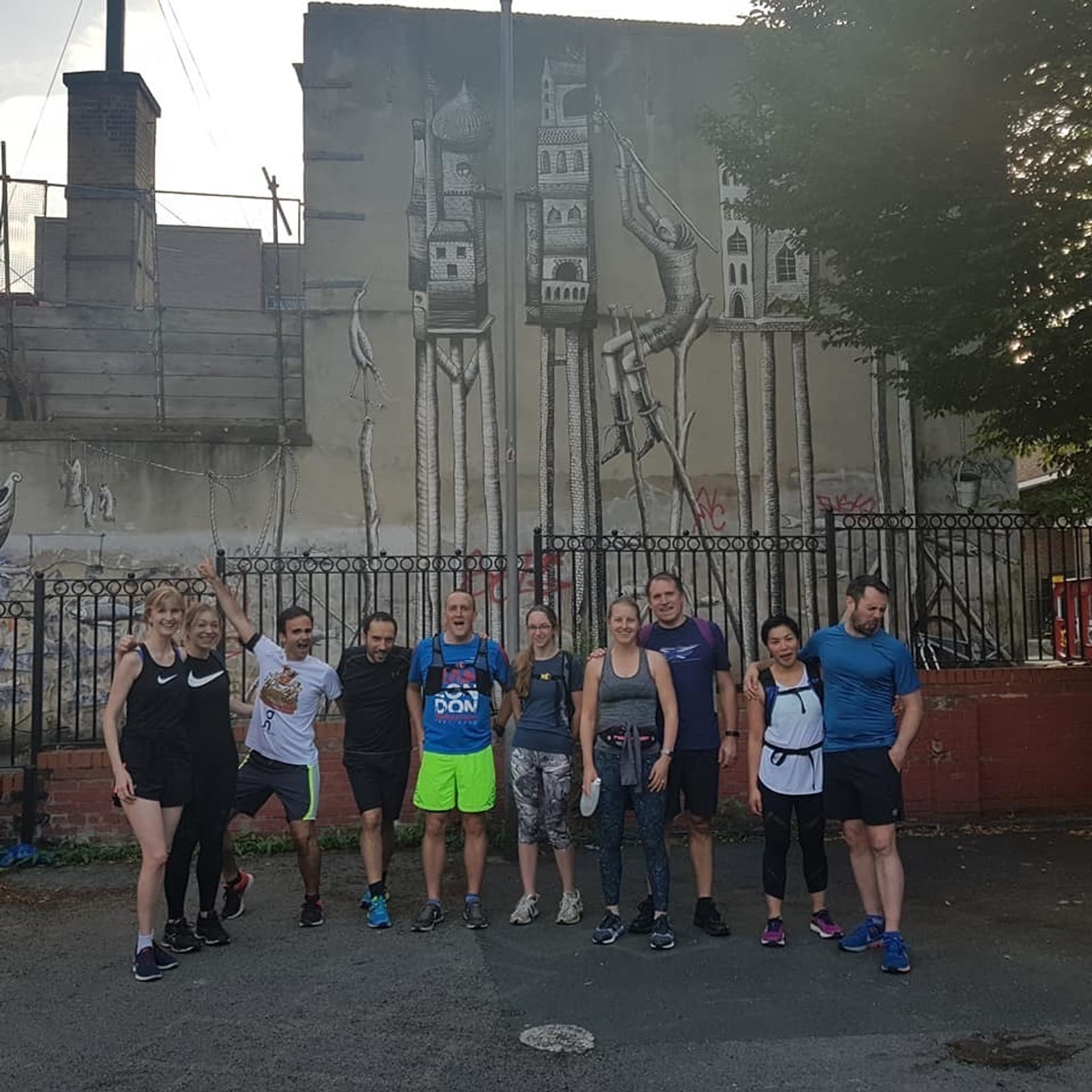 A group of ten people in athletic wear standing in front of a wall with street art. The art depicts tall, skinny buildings on stilts and plant-like designs. The group appears to be ready for or finishing a run, and they are posing for a photo with smiles and relaxed postures.