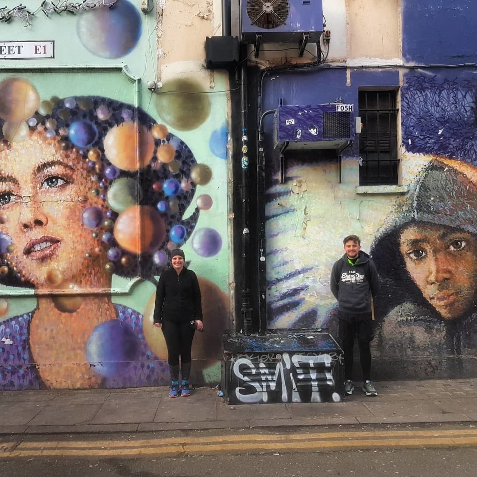 Two people stand in front of a wall with vibrant street art. On the left, a mural of a woman's face surrounded by colorful bubbles. On the right, a mural depicting a person in a hooded jacket. Both individuals smile and pose between the murals.