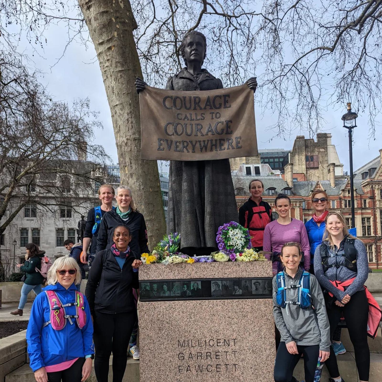 A group of ten people, wearing athletic clothing, gather around a statue of Millicent Garrett Fawcett holding a banner that reads "Courage calls to courage everywhere." The statue has flowers at its base and is located in an urban area with buildings and trees.
