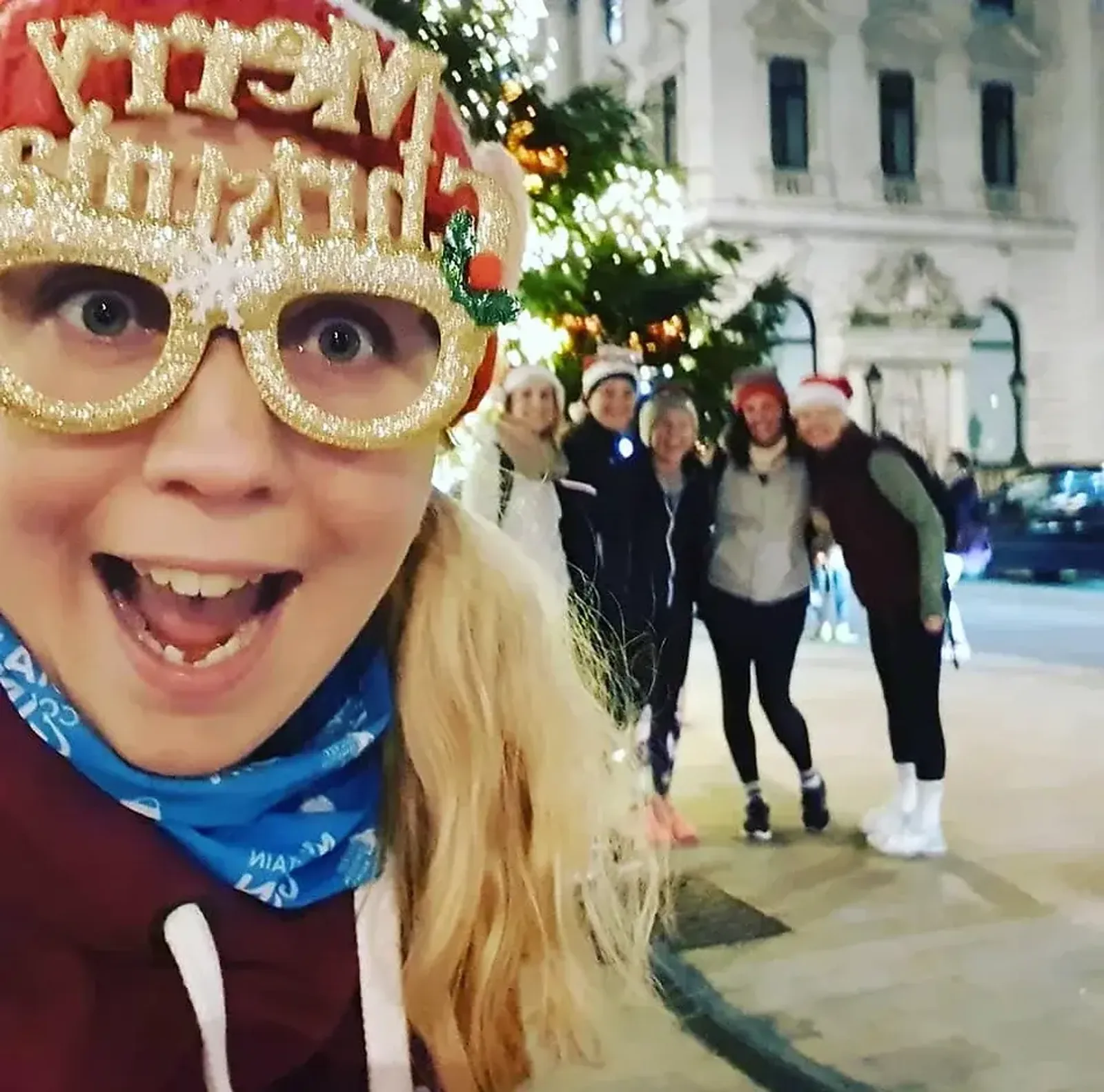 A joyful person with long blond hair is wearing glittery "Merry Christmas" glasses and a blue neck warmer, taking a selfie. Five other people, blurred in the background, are in festive street attire near a decorated Christmas tree in an urban setting.