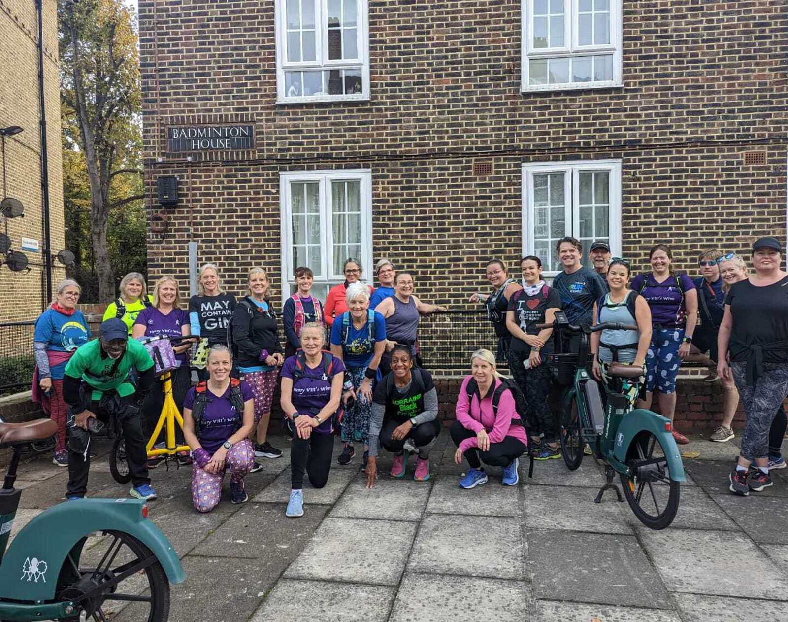 A group of diverse people, standing and smiling outside a brick building labeled "Badminton House." Some are in athletic wear and hold bicycles, suggesting they may have participated in an outdoor event or ride. The ground is paved and trees are visible in the background.