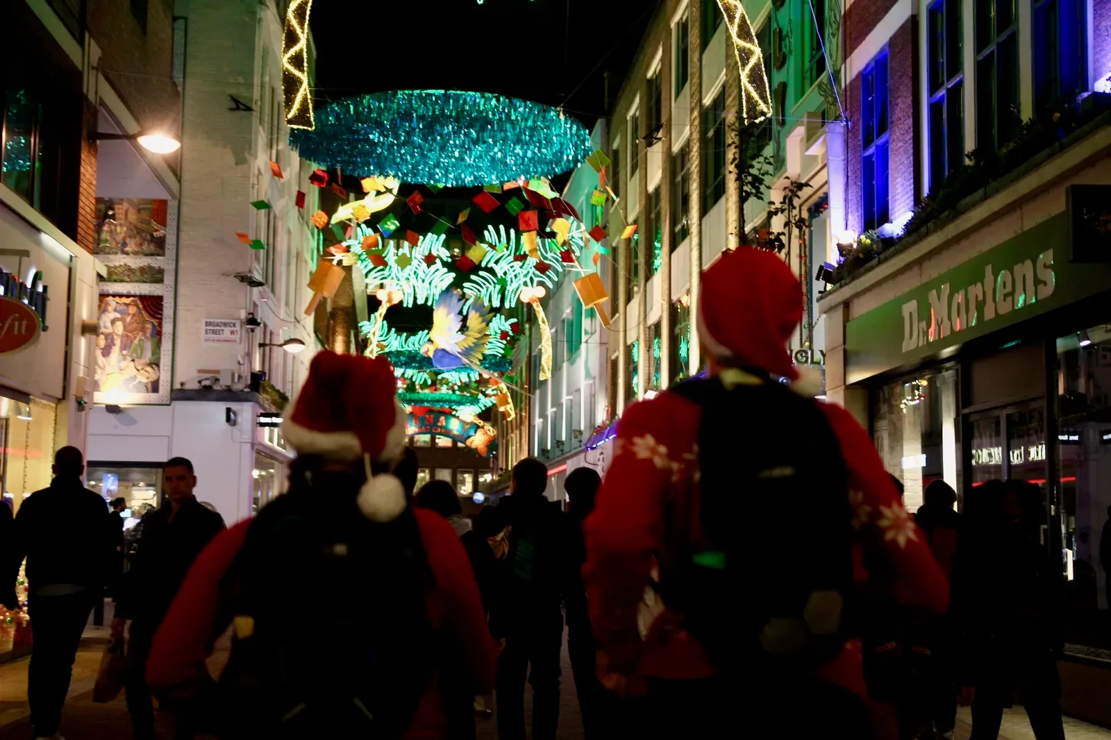 A festive street scene at night featuring colorful holiday lights and decorations hanging above a busy shopping street. People, including two individuals in Santa hats and red sweaters, are walking and enjoying the vibrant atmosphere.