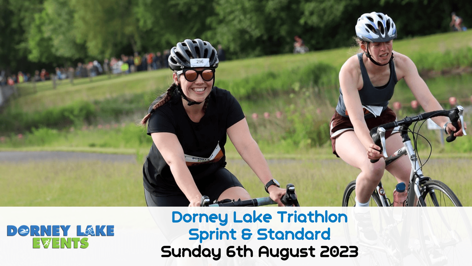Two cyclists, one in black and the other in blue, ride their bikes in an outdoor event on a sunny day. Both are wearing helmets and sunglasses. A banner reads "Dorney Lake Triathlon Sprint & Standard, Sunday 6th August 2023." The background shows greenery and participants.