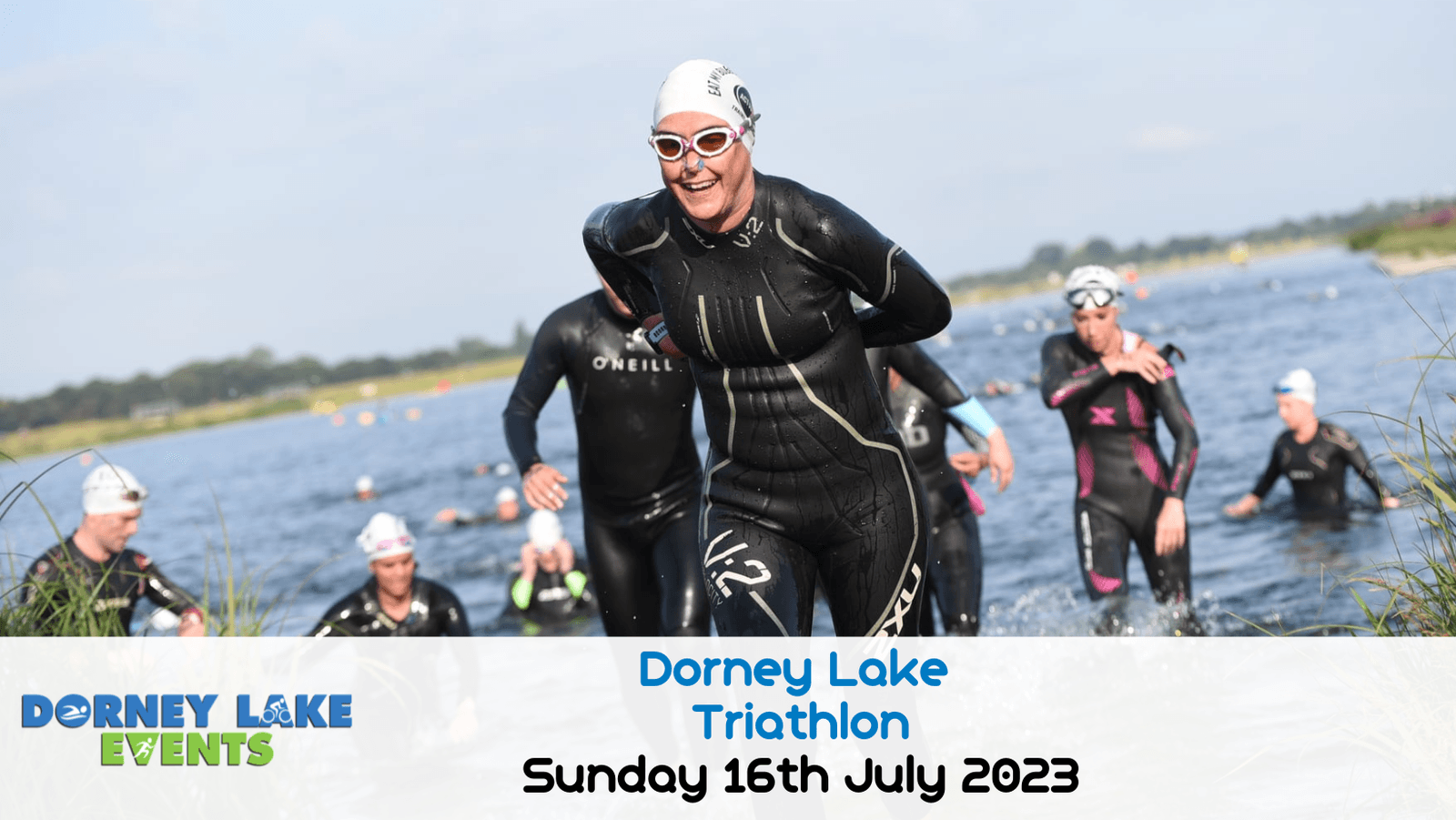 A group of triathletes in wetsuits, including a smiling front runner, emerge from the water during the Dorney Lake Triathlon. Text at the bottom reads "Dorney Lake Triathlon Sunday 16th July 2023". The event banner is in the lower left corner.
