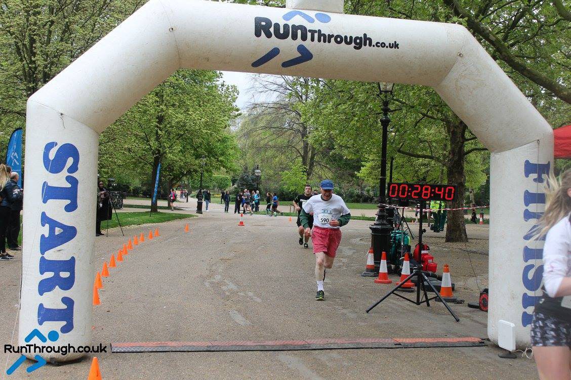 A runner wearing a white shirt and pink shorts crosses the finish line under an inflatable archway labeled "START" and "FINISH." The timer shows 28:40. Other participants can be seen running in the background on a path lined with trees.