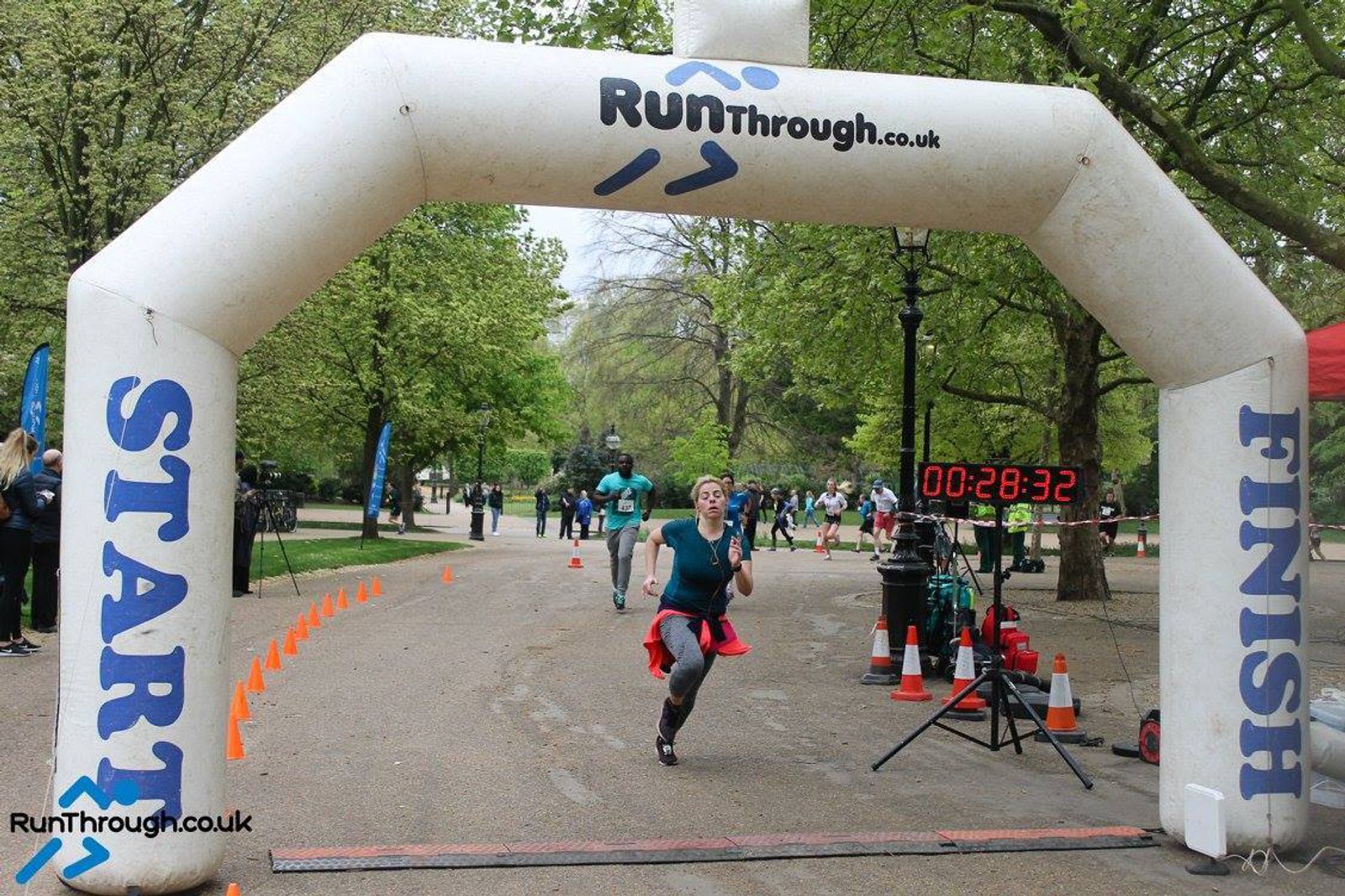 A woman is running through the finish line under an inflatable archway labeled "START" on one side and "FINISH" on the other. A digital timer displays 28:32. She is in a park setting with other runners and trees in the background. "RunThrough.co.uk" is printed on the arch.