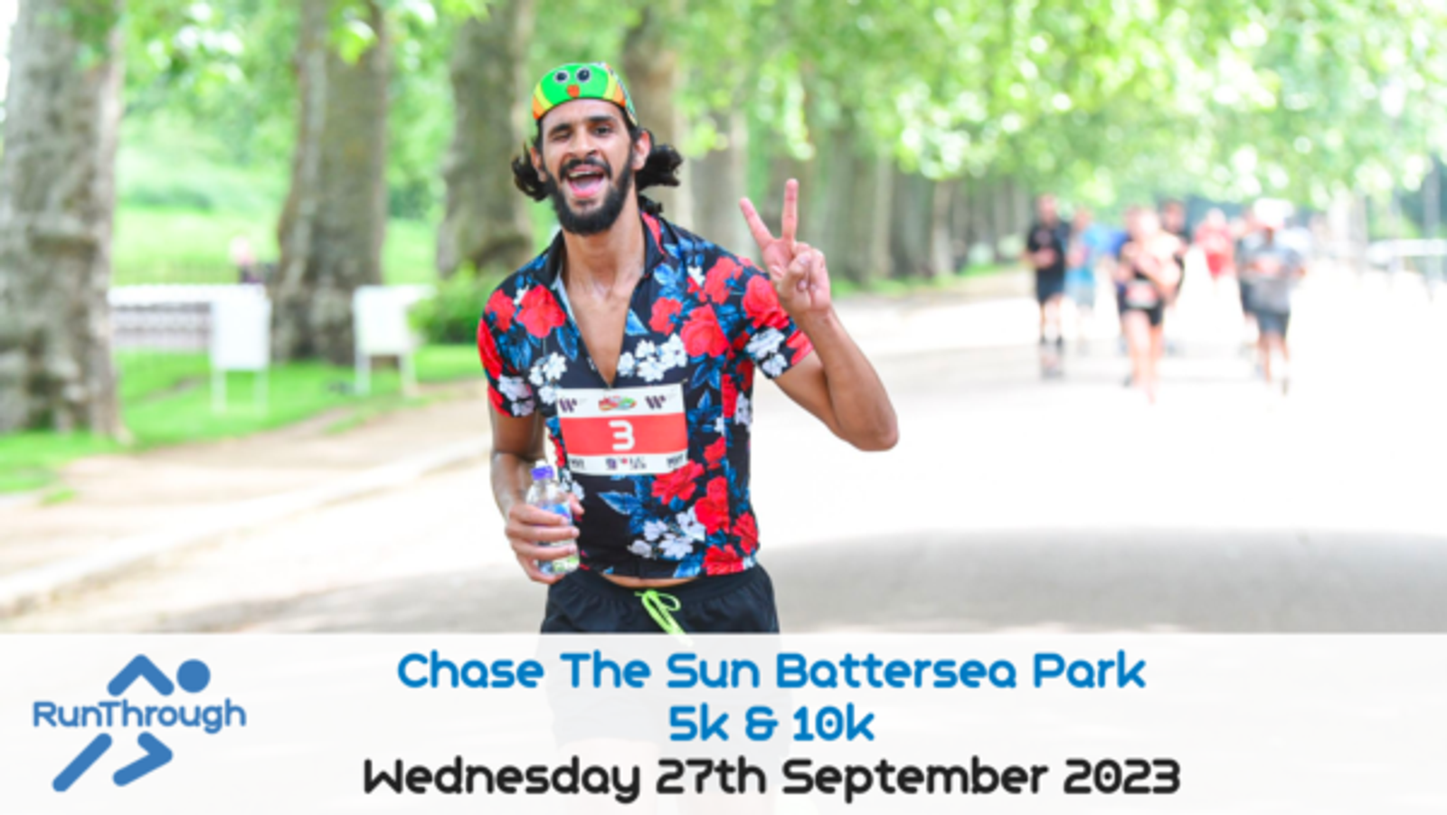 A male runner wearing a floral shirt, a cardboard frog crown, and race bib number 3, flashes a peace sign while running in Chase The Sun Battersea Park 5K & 10K on Wednesday 27th September 2023. Other runners and trees line the path behind him. RunThrough logo displayed.