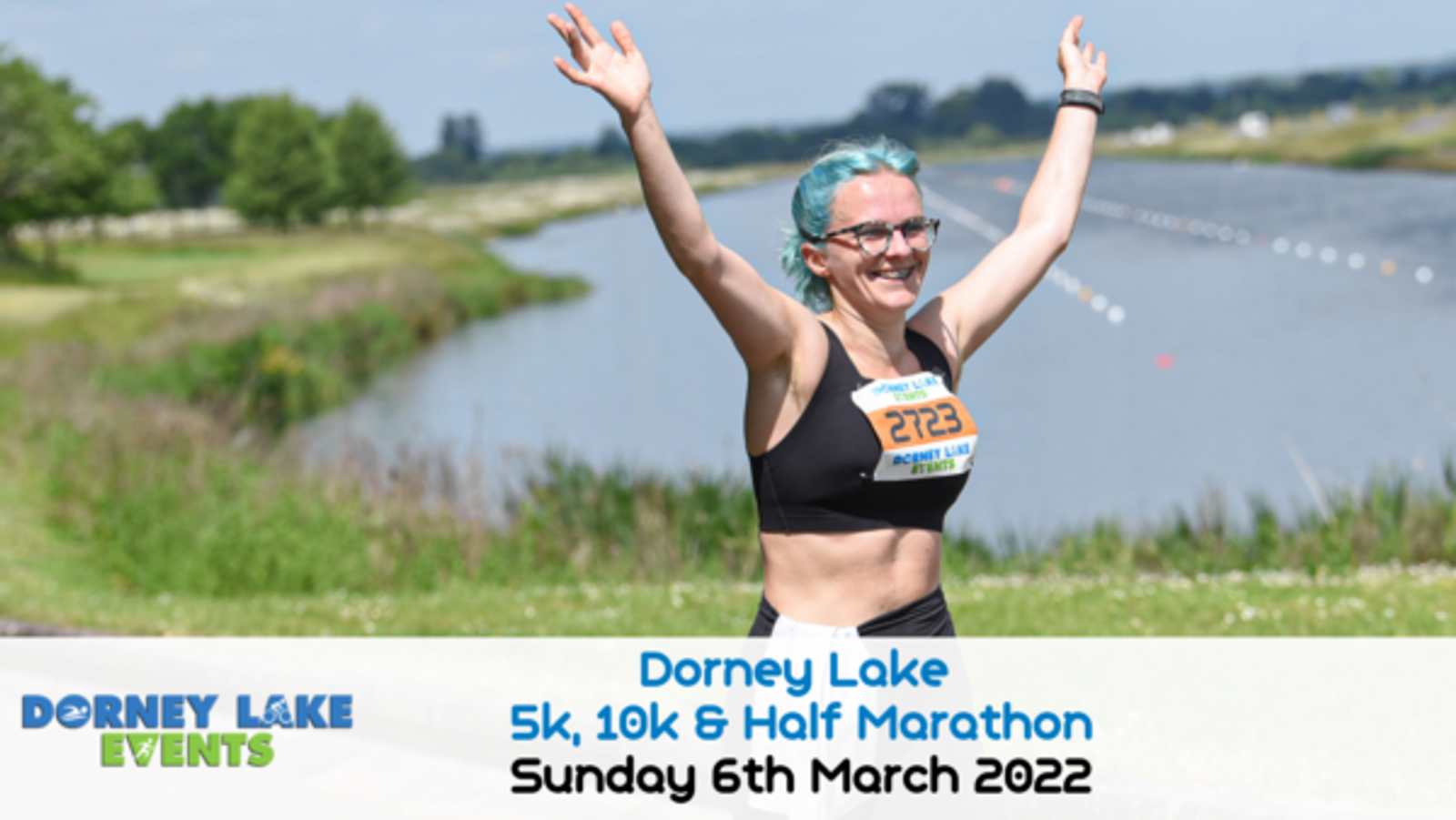 A runner with blue hair and glasses smiles with arms raised in victory as they cross the finish line at an outdoor race beside a lake. The banner at the bottom reads, "Dorney Lake 5k, 10k & Half Marathon, Sunday 6th March 2022.
