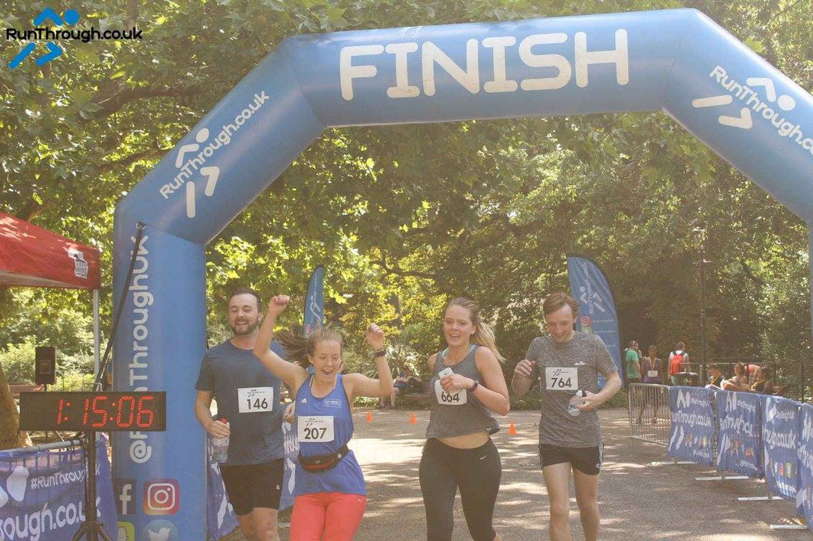Four runners celebrate as they cross the finish line of a race. They are under a blue inflatable arch with "FINISH" written on it. The timing clock reads 1:15:06. The background shows more participants and lush trees. All runners are smiling and appear jubilant.