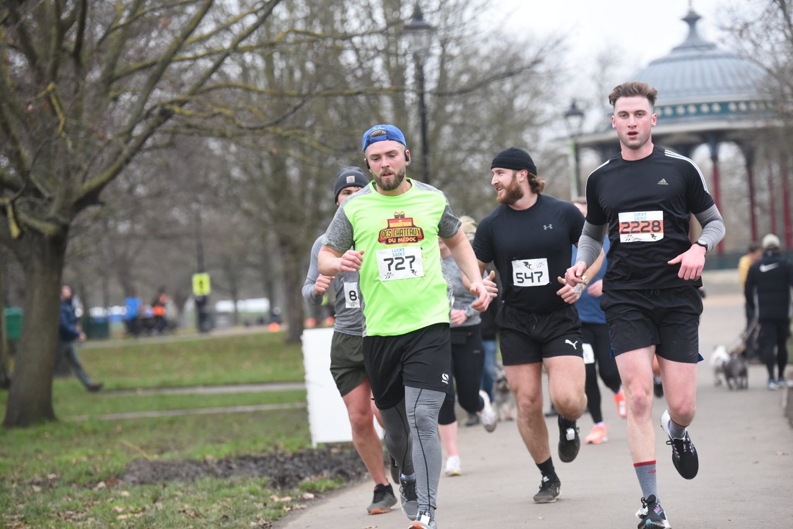 Runners participating in a race in a park. The focus is on several men wearing numbered bibs, with one wearing a bright green shirt and blue cap. Trees and a gazebo are in the background, and the path is lined with spectators.